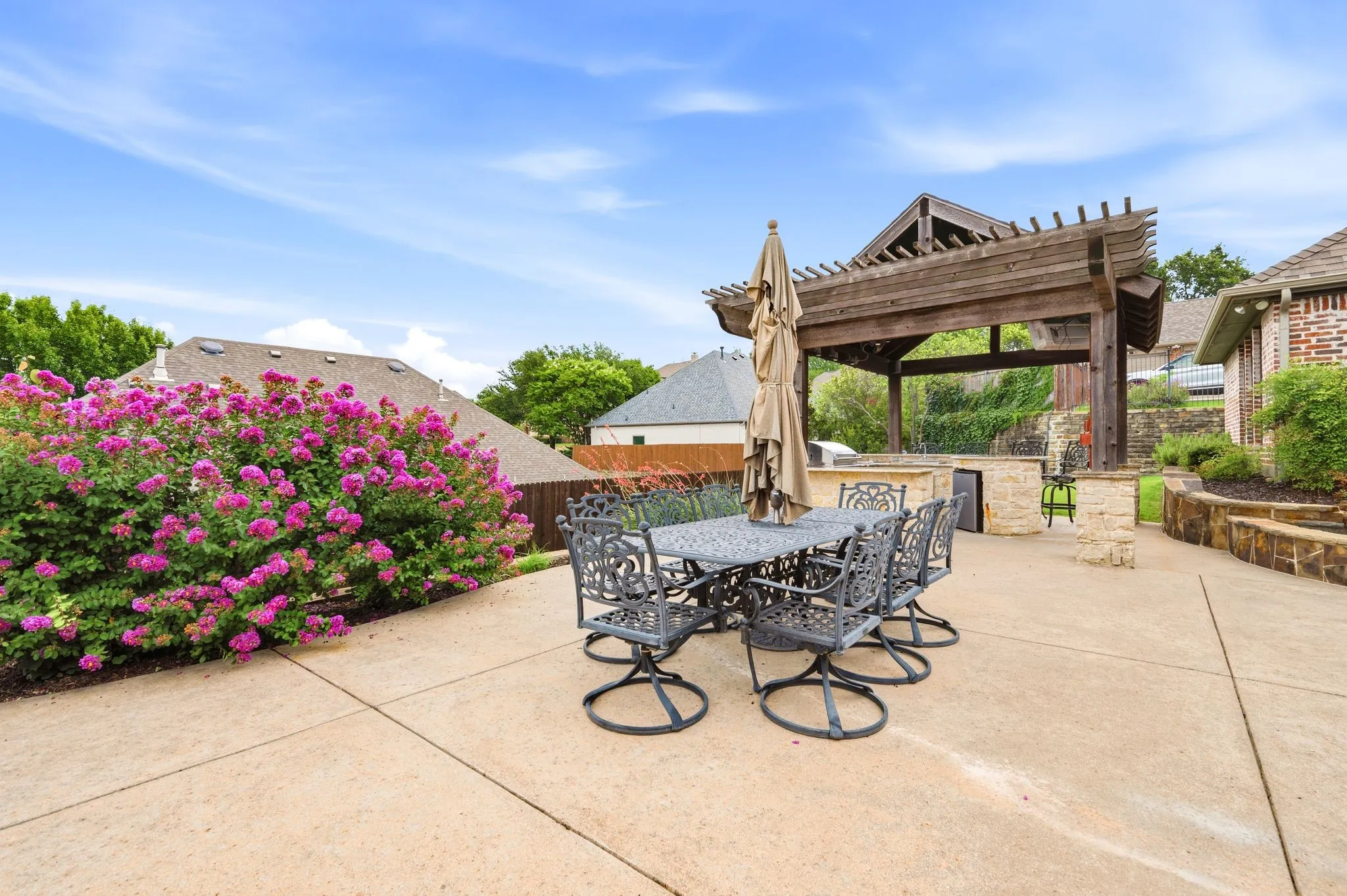 Expansive outdoor patio featuring a large wooden pergola, built-in stone outdoor kitchen, stone retaining walls, and vibrant flowering shrubs