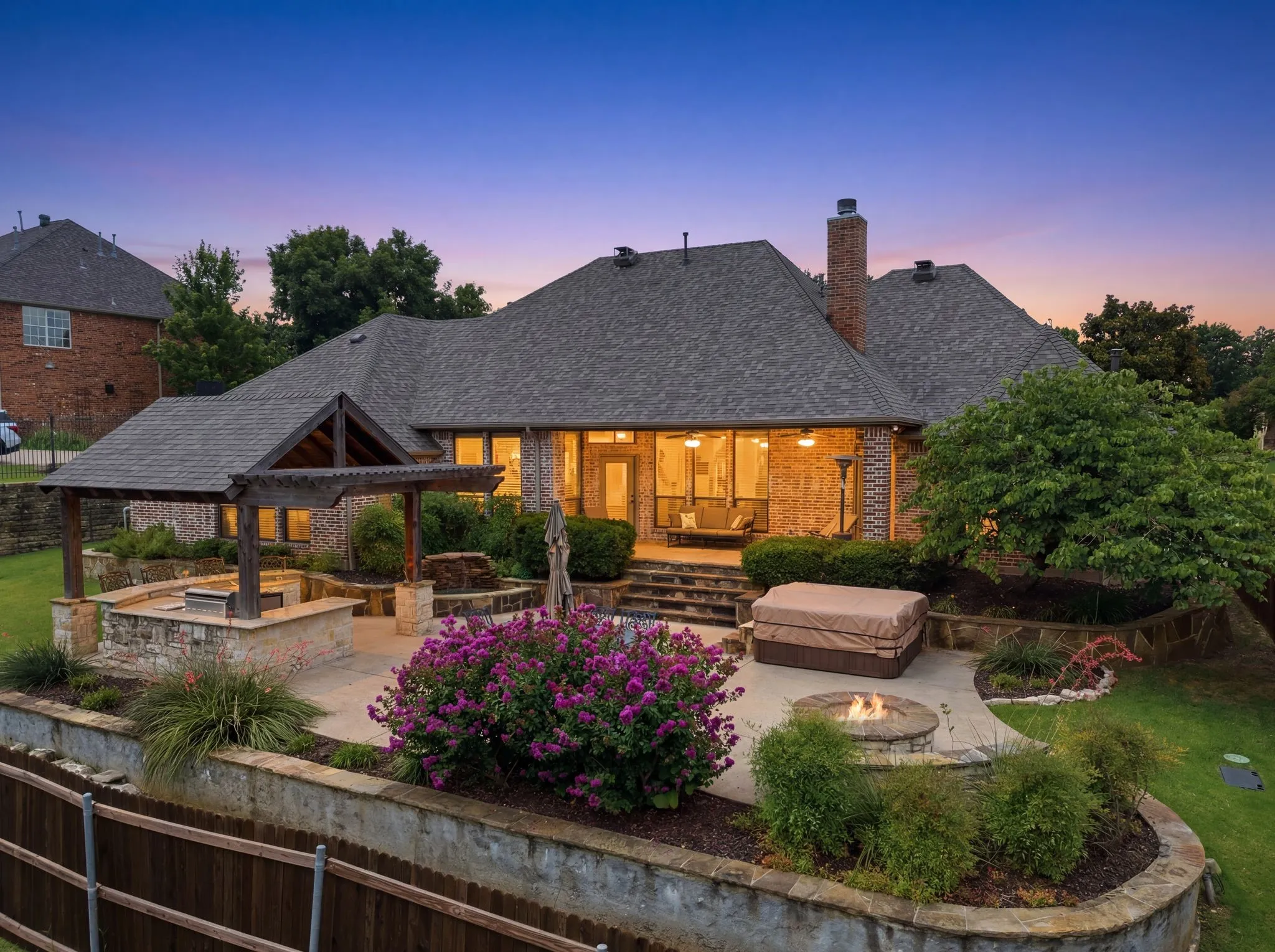 Expansive backyard featuring a multi-level stone patio, a covered outdoor kitchen area with built-in grill, a circular stone fire pit, and mature landscaping with flowering shrubs