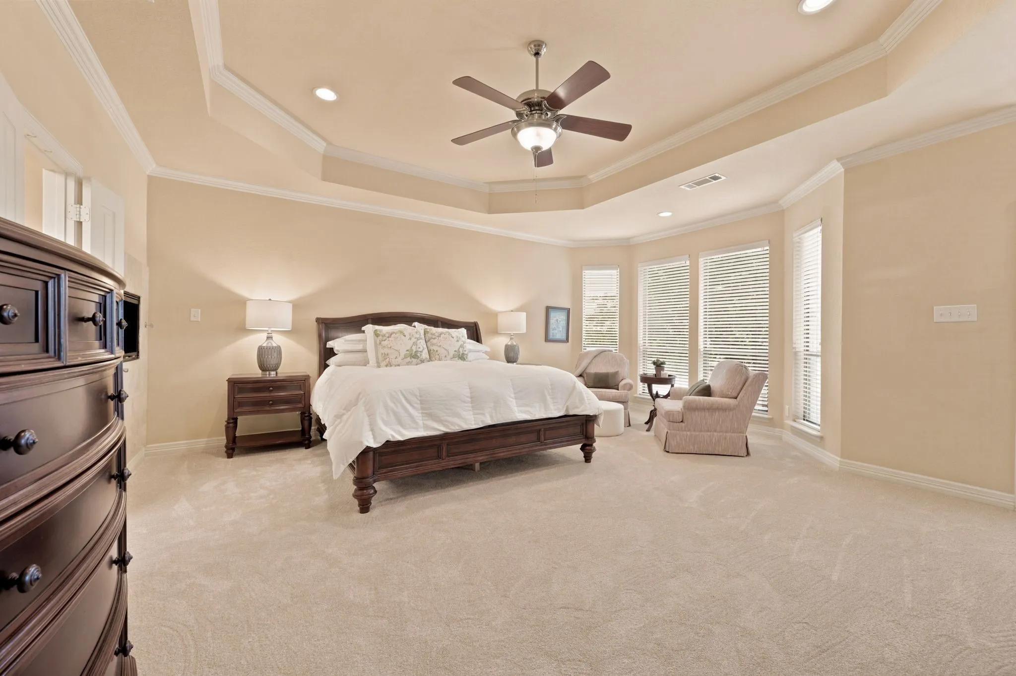 Spacious bedroom featuring plush carpeting, a tray ceiling with recessed lighting and a ceiling fan, and a bay window area with multiple windows