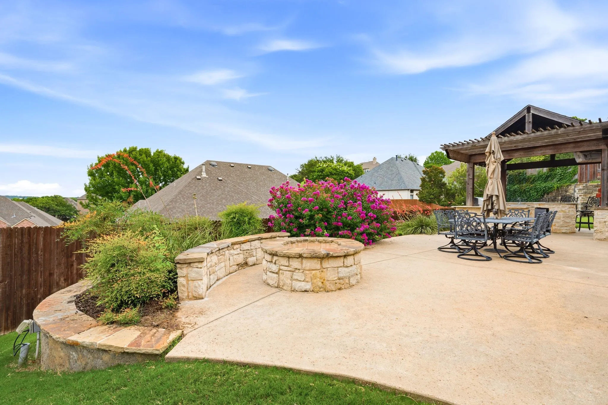 Expansive concrete patio featuring a stone-clad fire pit, a curved stone retaining wall with integrated planting beds, and a wood pergola structure