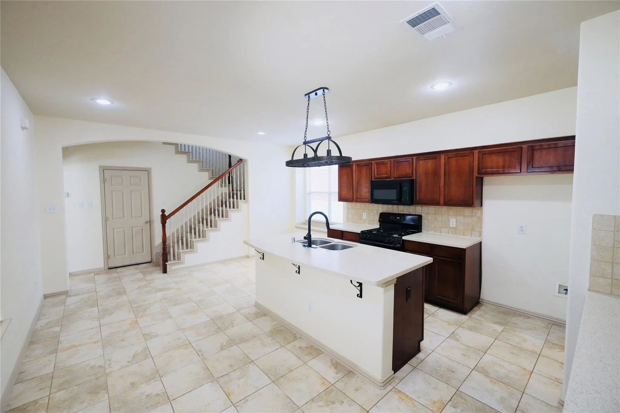 Tiled flooring throughout, kitchen island with built-in sink and seating, dark wood-finish cabinetry, tile backsplash, and a staircase with a wood-finish handrail and balusters