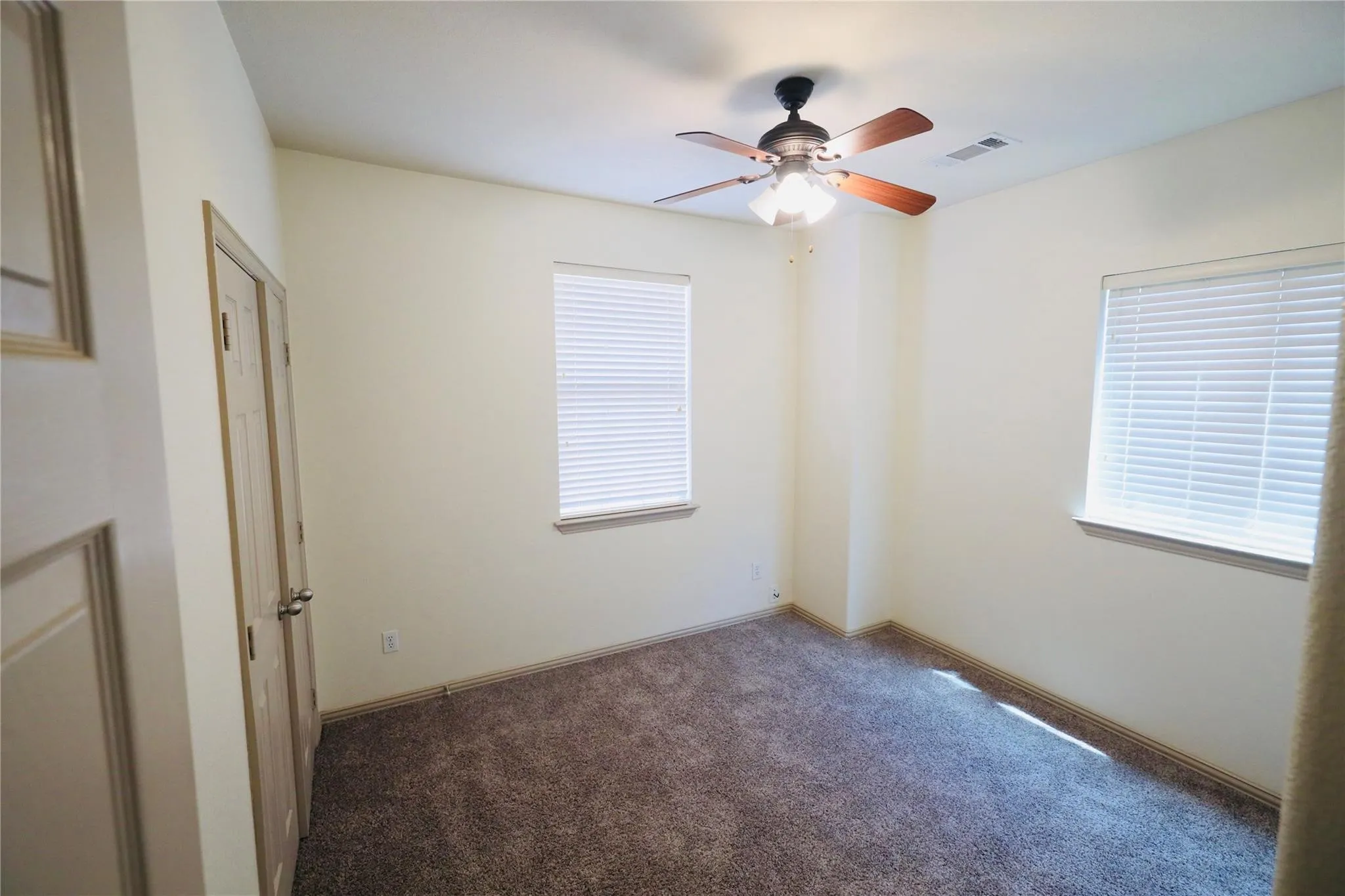 Ceiling fan with light fixture, two windows with blinds, light-toned walls, and carpet flooring