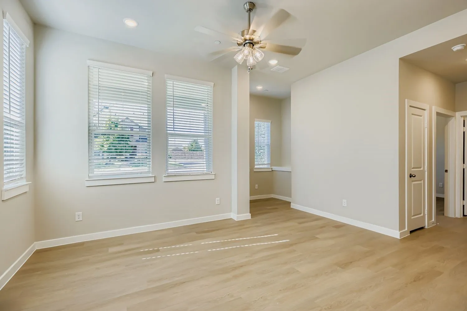 Neutral-toned interior space featuring wood-finish flooring, recessed lighting, a brushed-metal ceiling fan with integrated glass lighting, and multiple windows with installed blinds