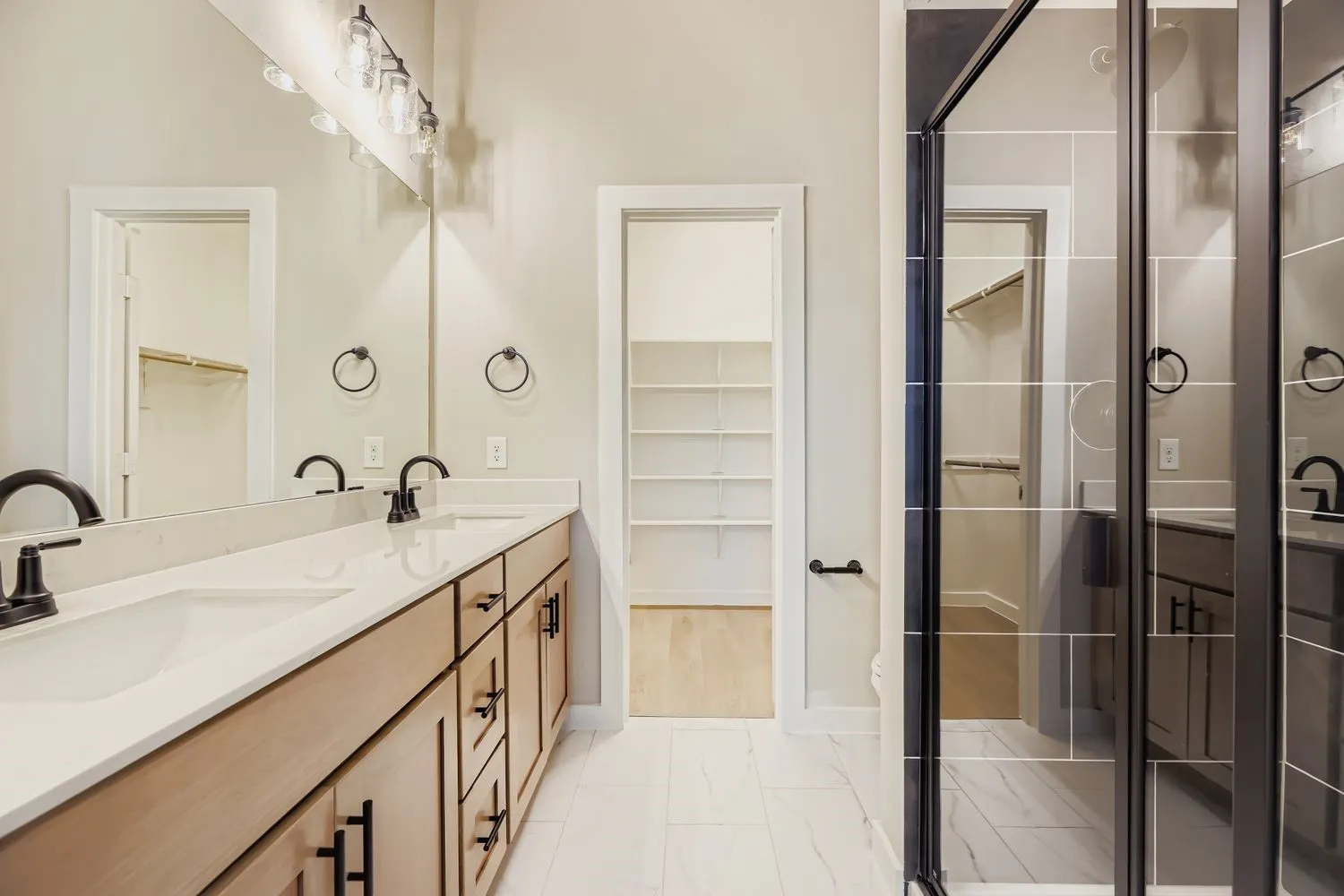 Dual-sink vanity with light wood-finish cabinetry, white solid-surface countertops, and matte black fixtures, paired with a glass-enclosed shower featuring large-format tile surround and a walk-in storage area with built-in shelving