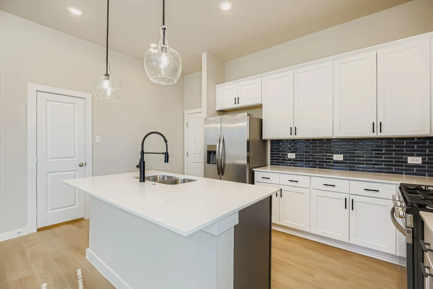 Kitchen featuring white shaker-style cabinetry, quartz countertops, a dark-toned subway tile backsplash, a central island with a double-basin sink and matte black faucet, and pendant lighting