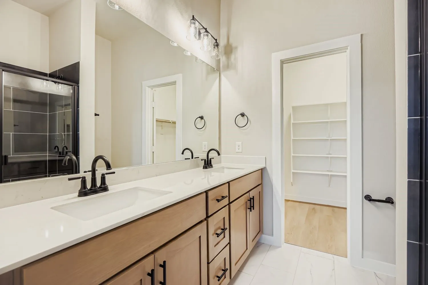 Dual-sink vanity with light-tone wood cabinetry, quartz countertops, matte black fixtures, and a wall-to-wall mirror