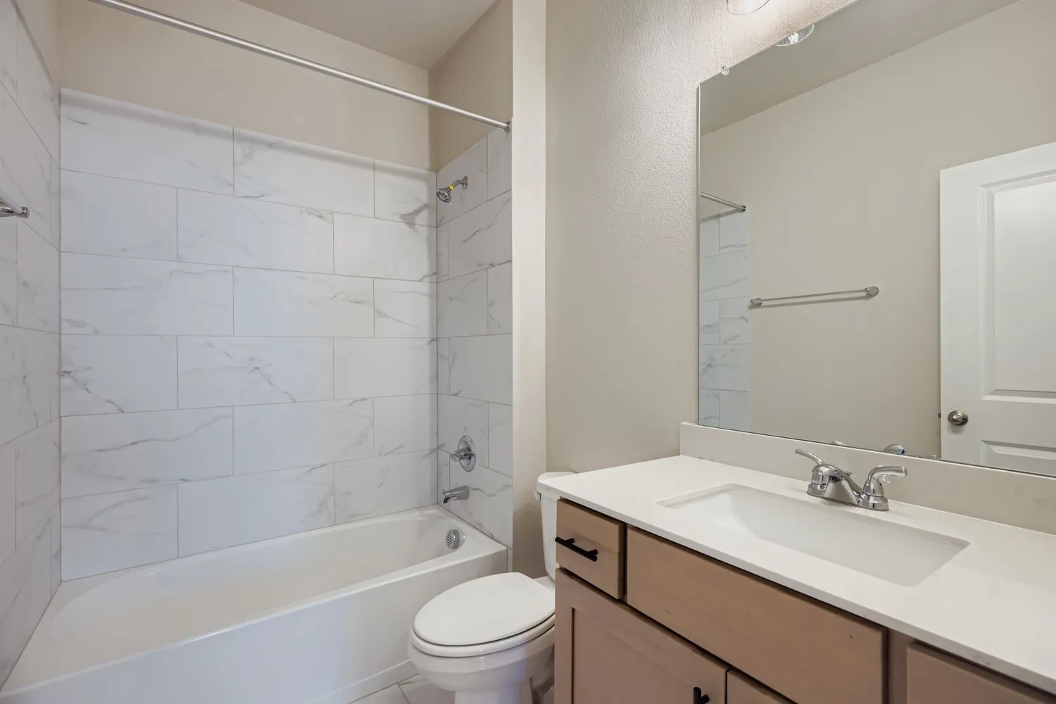 Bathroom featuring a vanity with a light-toned wood finish, white countertop, integrated sink, and chrome fixtures, paired with a shower-tub combo enclosed by white tile with a stone-pattern finish and a full-width vanity mirror