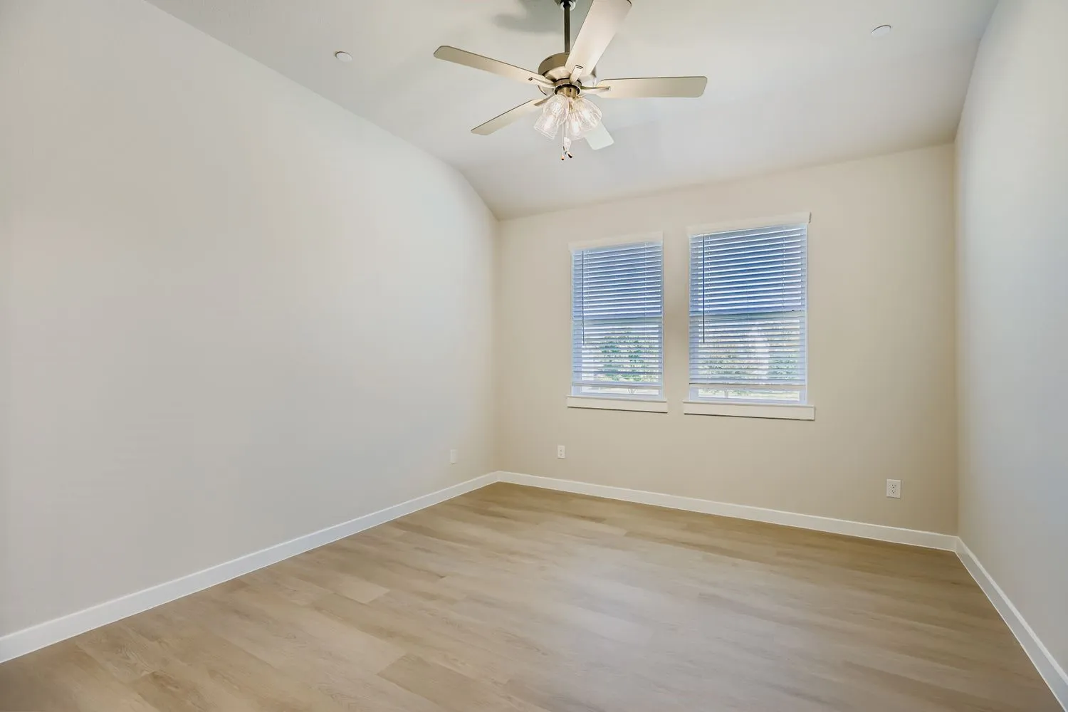 Room with vaulted ceiling, neutral wall tones, wood-finish flooring, dual windows with blinds, and a central ceiling fan with integrated light fixture