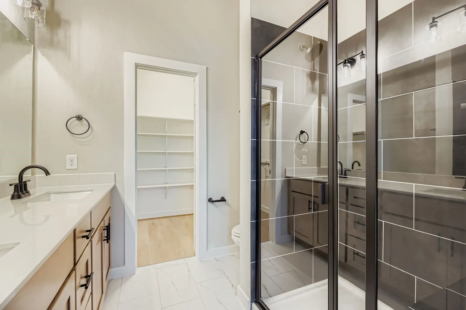 Dual-sink vanity with light-toned cabinetry and quartz countertops, walk-in shower with dark-toned tile surround and matte black hardware, and built-in shelving