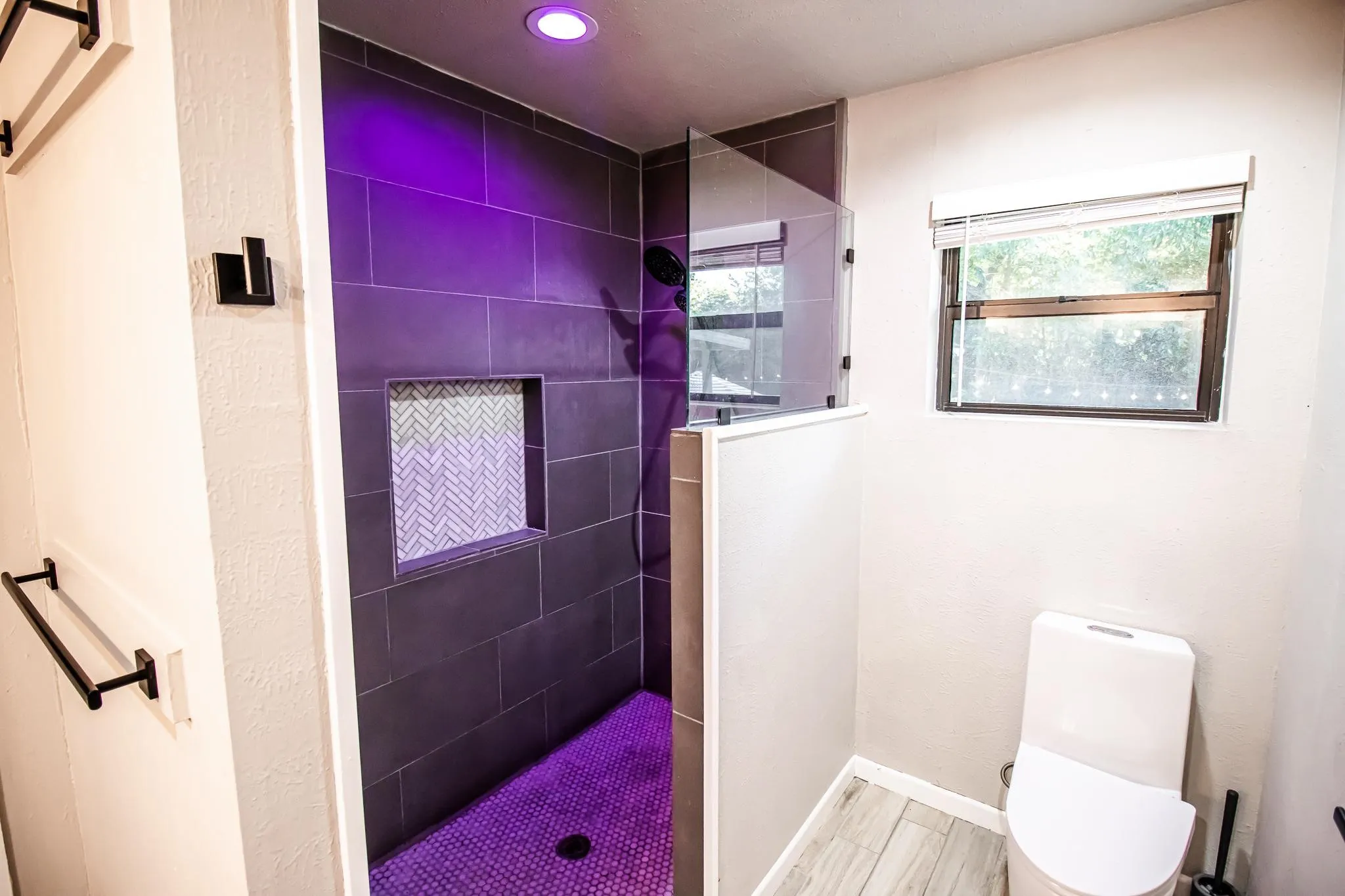 Walk-in shower featuring large-format dark tile, integrated herringbone-patterned wall niche, frameless glass panel, and matte black fixtures, complemented by wood-finish flooring