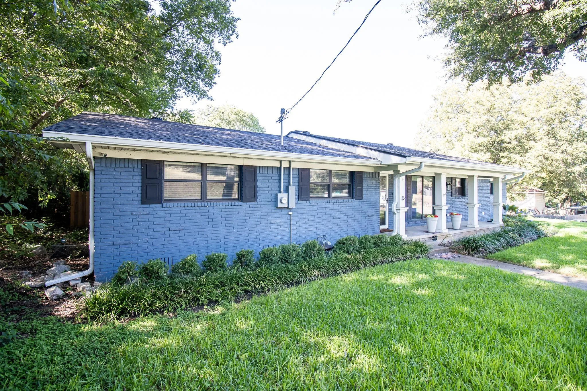 Single-story residence featuring blue painted brick exterior, covered front porch with square columns, and mature landscaping