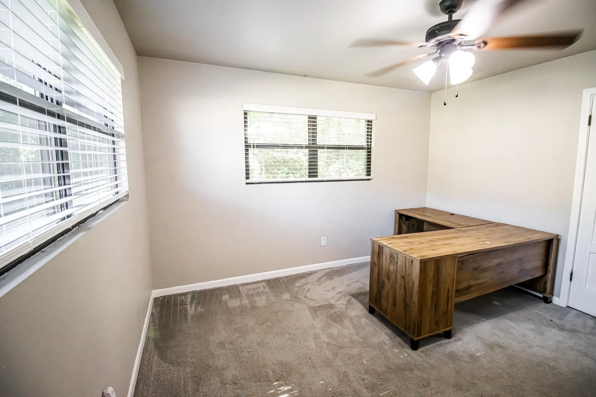 Neutral-toned room featuring neutral wall-to-wall carpeting, dual windows with horizontal blinds, and a ceiling-mounted light fixture with fan
