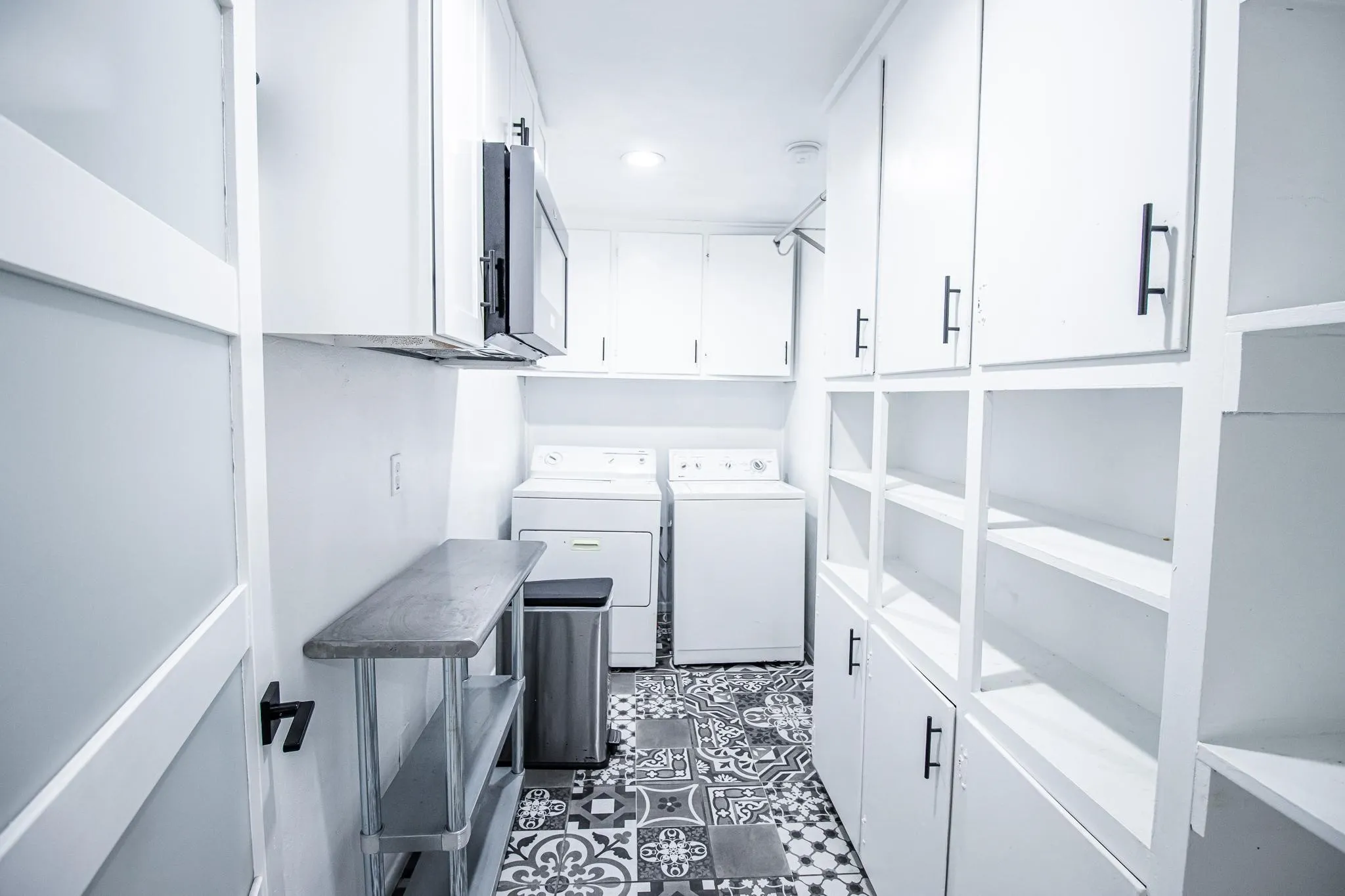 Laundry area featuring white cabinetry with matte black hardware, patterned tile flooring, an over-the-range microwave, and built-in open shelving