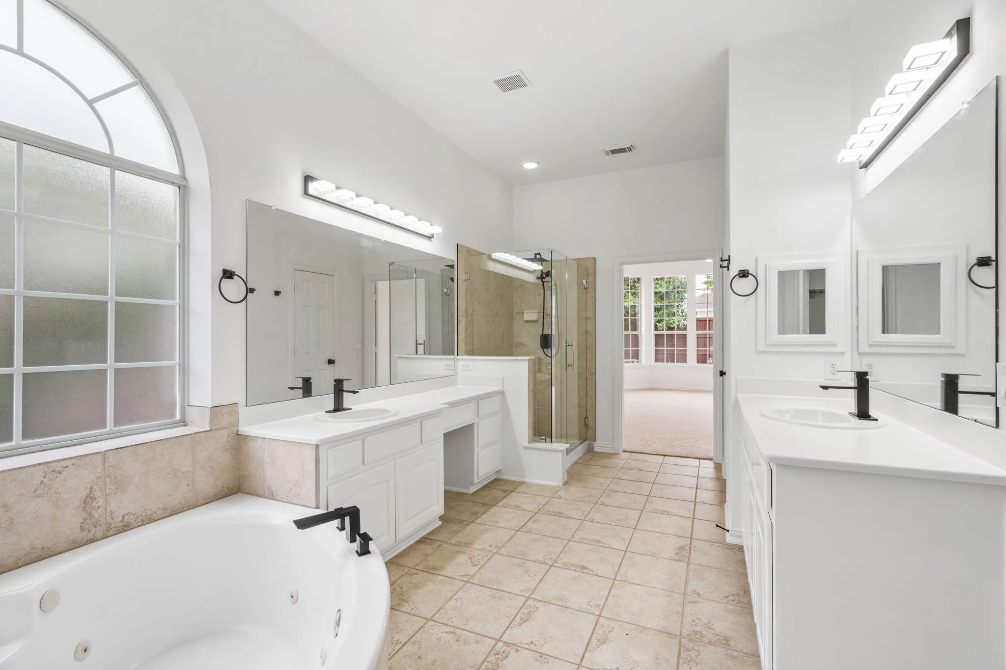Spacious bathroom featuring a jetted soaking tub, walk-in shower with glass enclosure, dual vanities with white cabinetry and integrated sinks, and an arched transom window