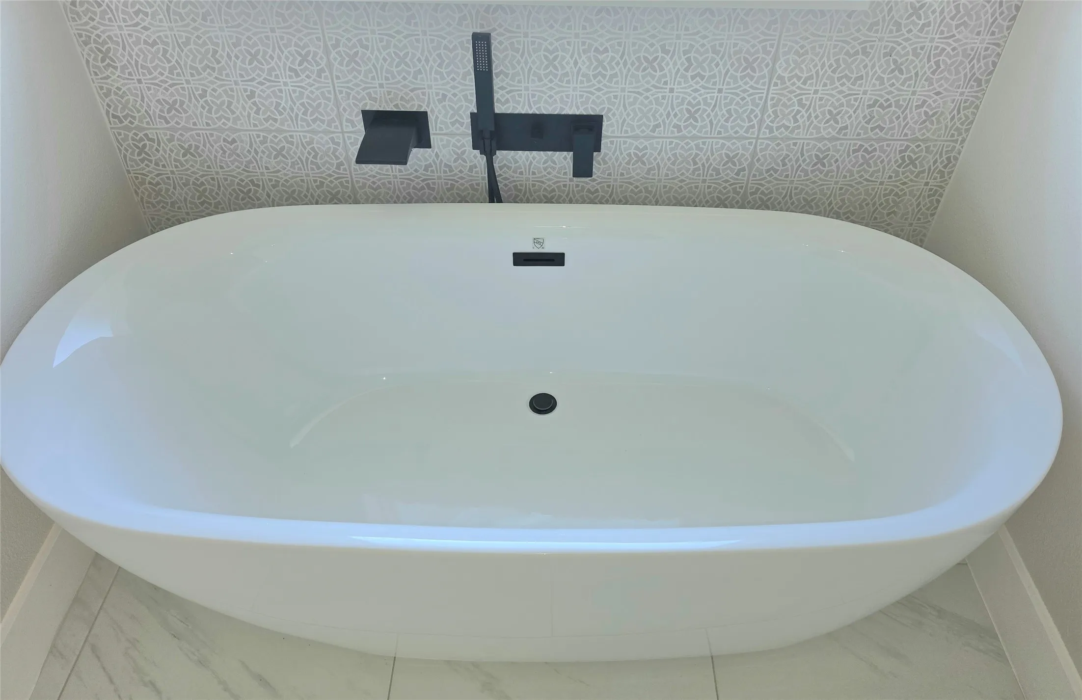 Freestanding soaking tub with matte black wall-mounted faucet and handheld shower attachment, accented by patterned wall tile and large-format floor tile