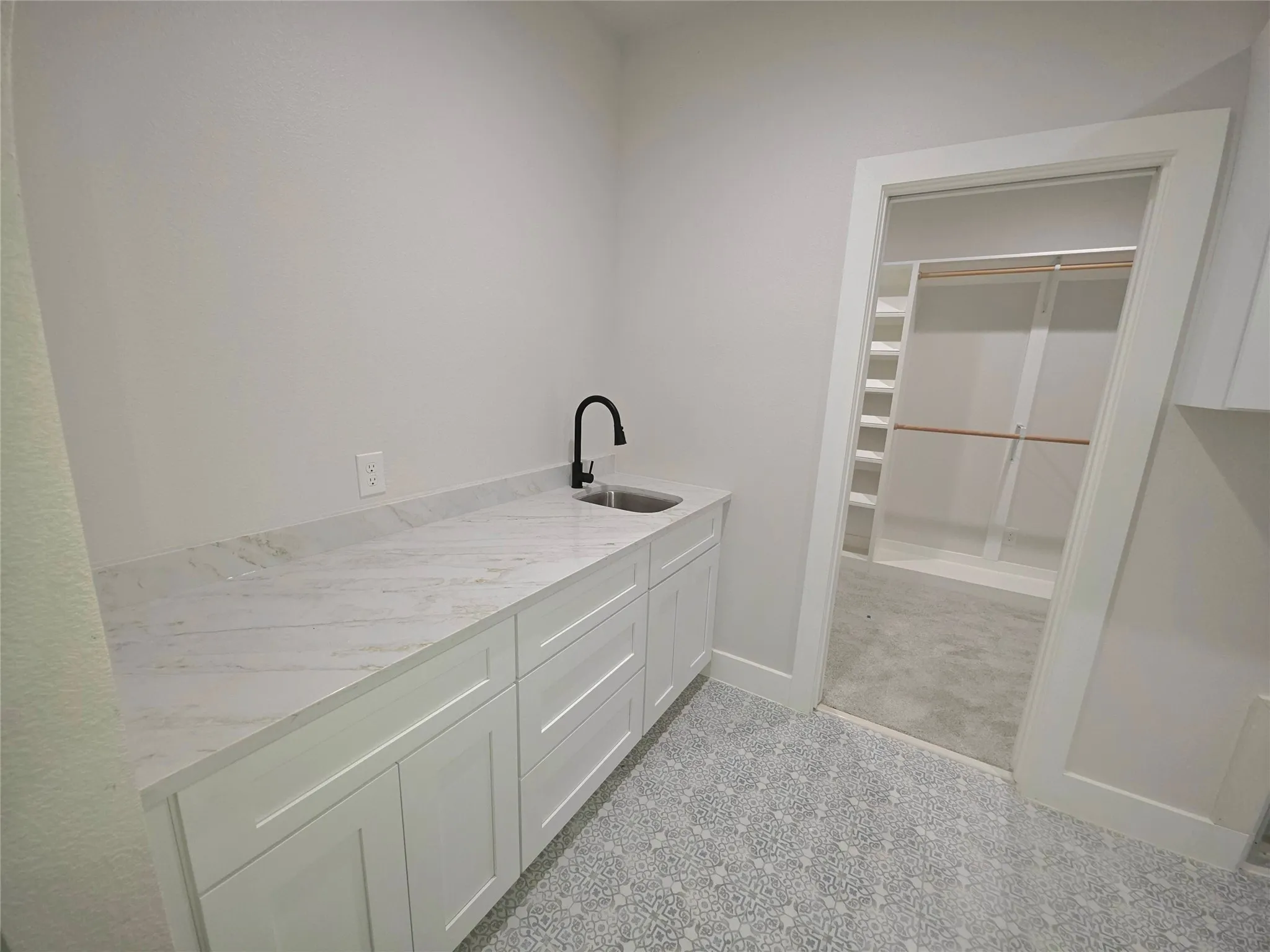 Utility space featuring white shaker-style cabinetry, stone-patterned countertops, a stainless steel undermount sink, and a matte black gooseneck faucet, with a doorway leading to a walk-in closet with built-in shelving