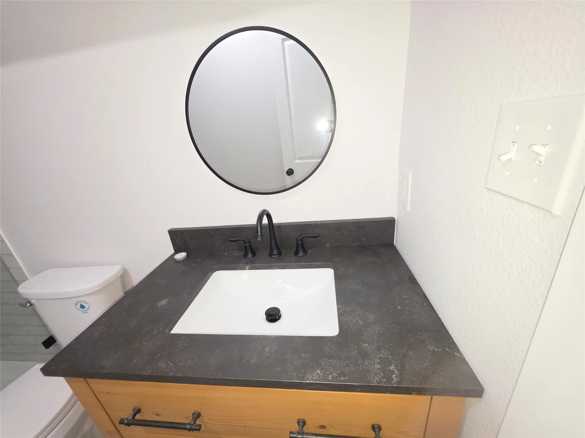 Vanity featuring a light wood finish, dark stone-style countertop, integrated rectangular sink, matte black widespread faucet, and a circular wall-mounted mirror