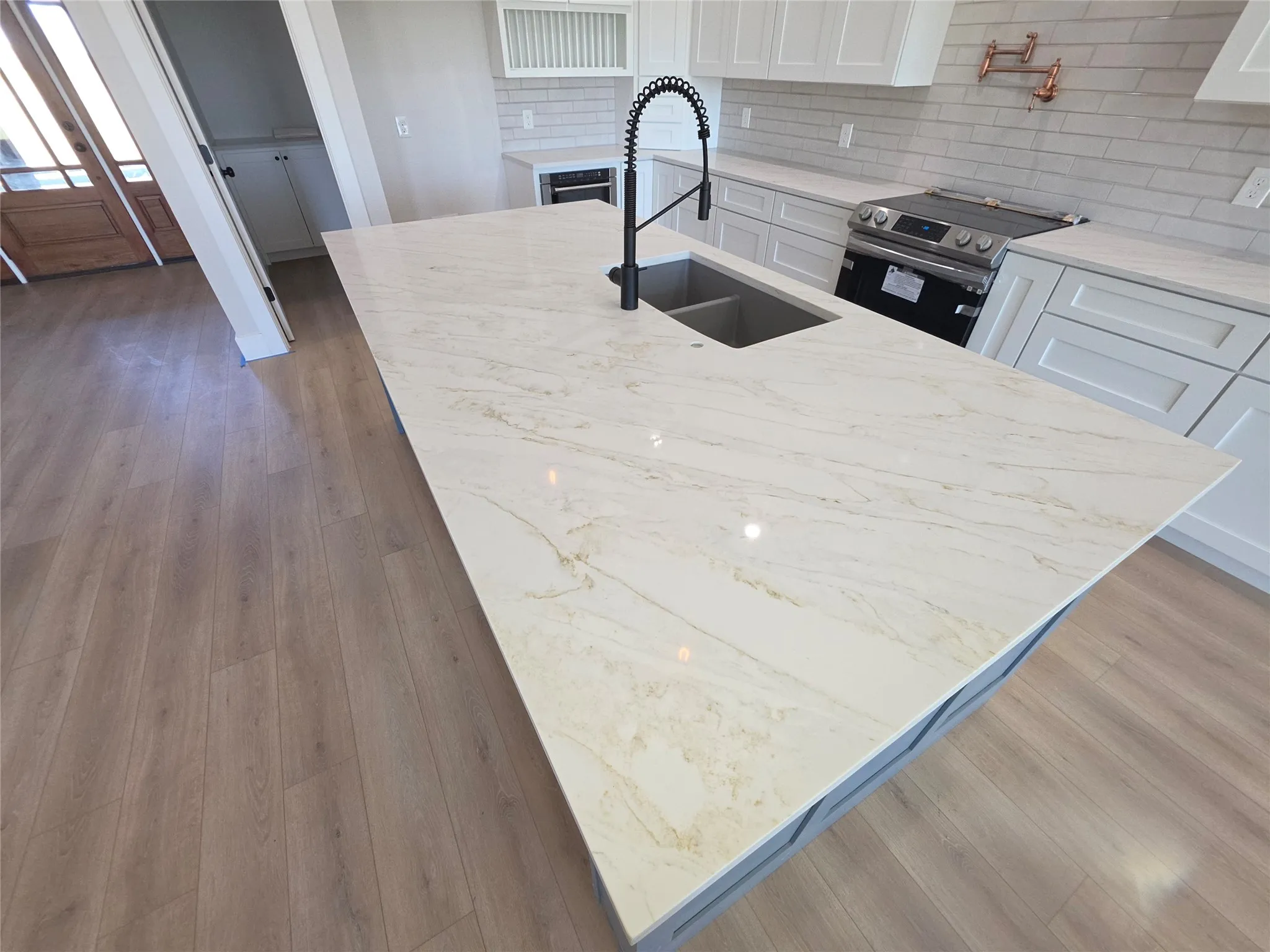Kitchen island featuring stone countertops, an undermount dual-basin sink with a high-arch pull-down faucet, white shaker-style cabinetry, a tile backsplash, and wood-finish flooring