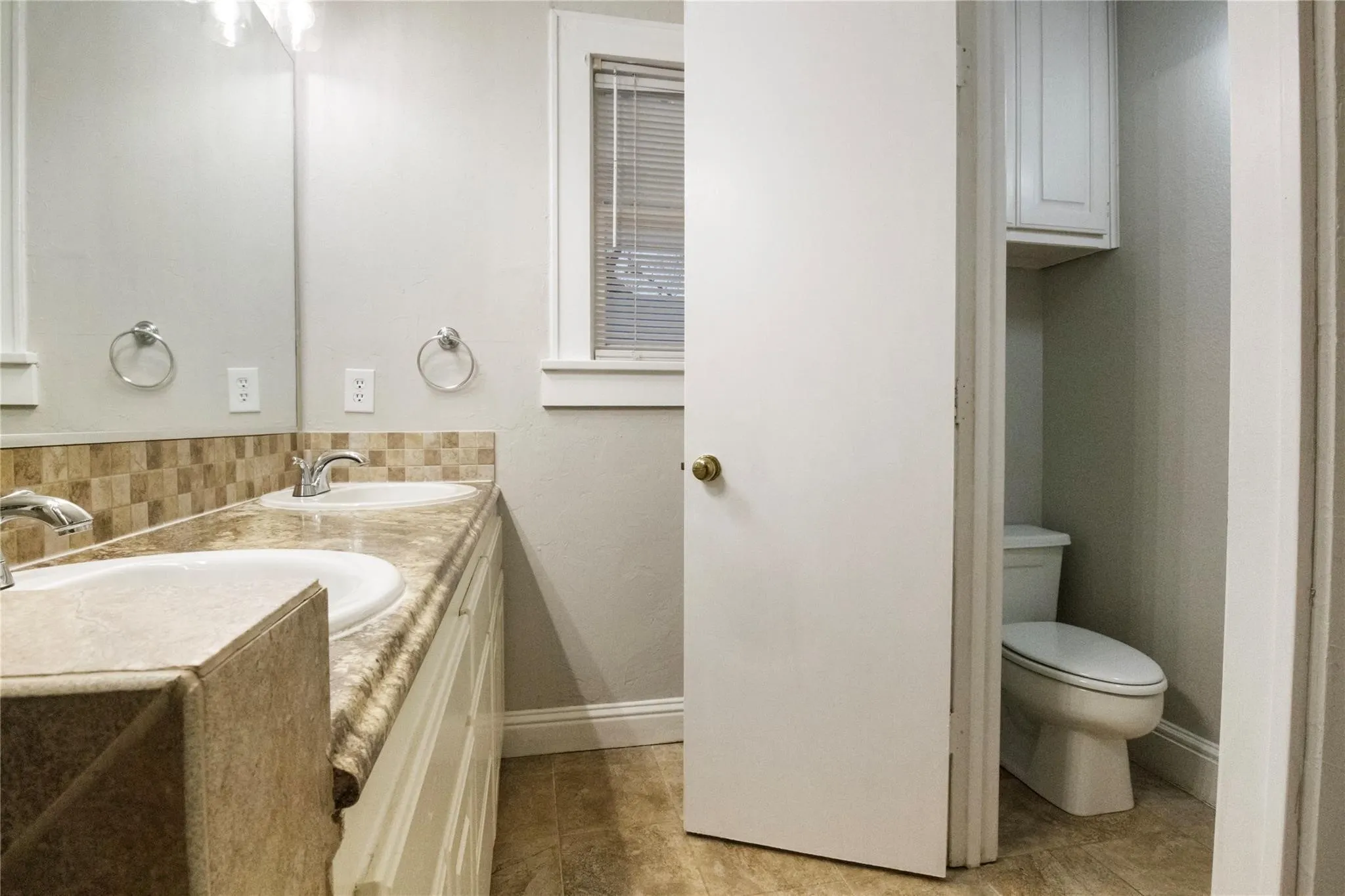 Dual-sink vanity featuring a stone-finish countertop, tiled backsplash, large wall-mounted mirror, and overhead cabinet storage