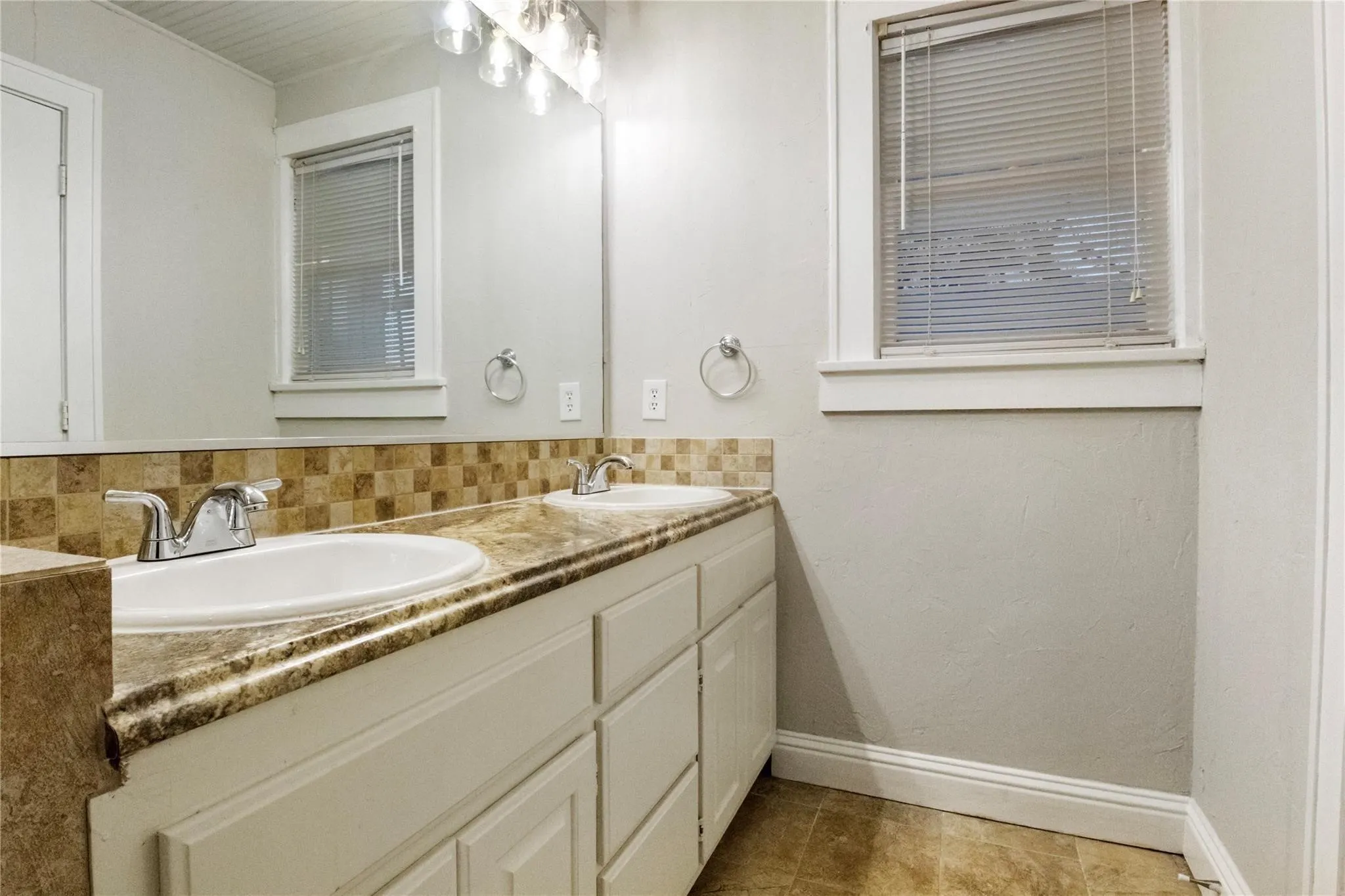 Dual-sink vanity featuring stone-finish countertops, tiled backsplash, white cabinetry, and polished chrome hardware