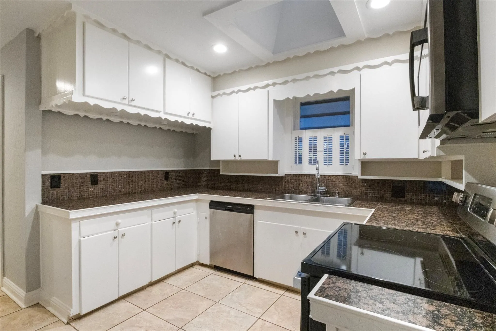 Kitchen featuring white cabinetry with scalloped valance detailing, stone-finish countertops, tiled backsplash, stainless steel dishwasher, and ceramic tile flooring
