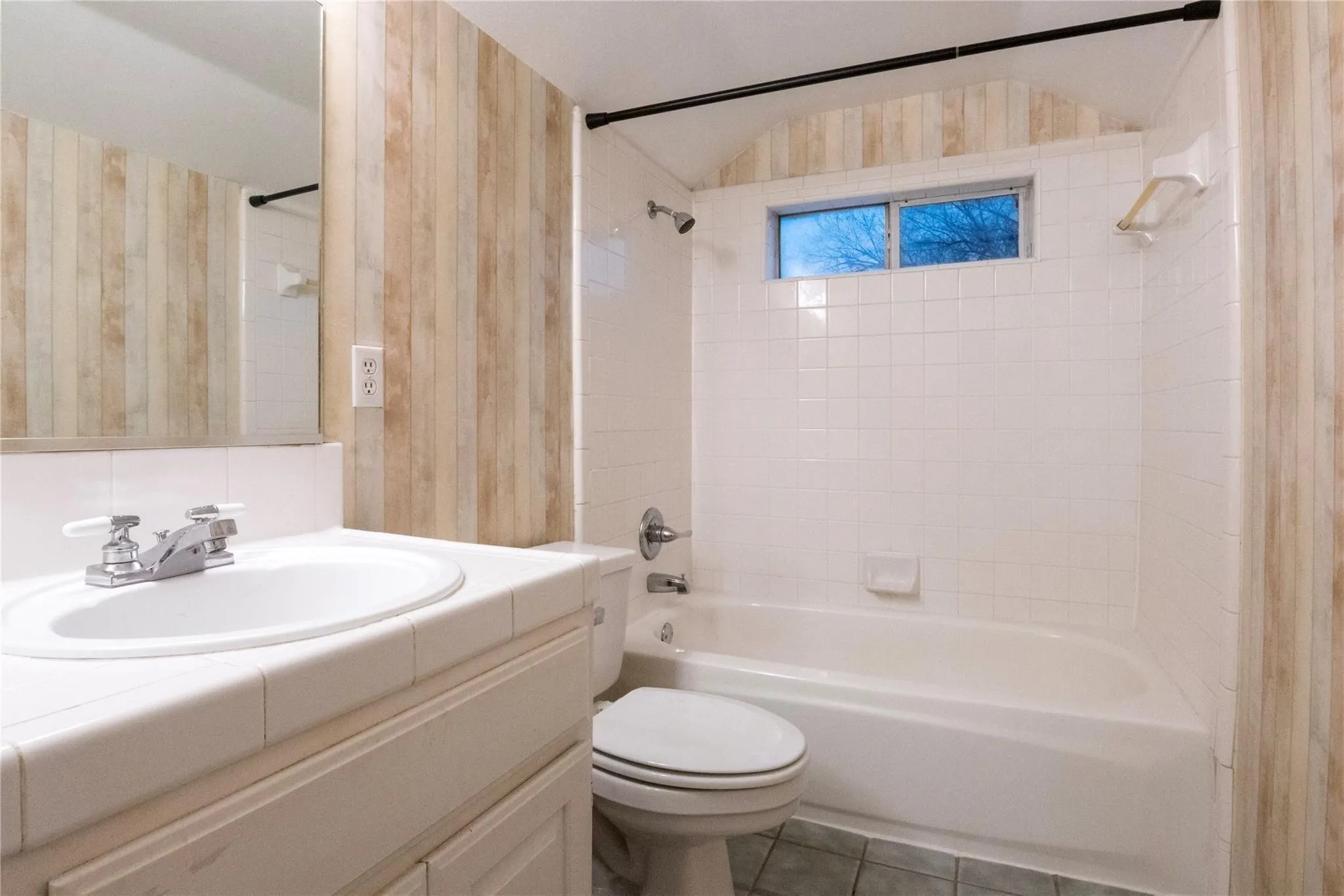 Bathroom featuring vertical wood-finish wall paneling, a white tiled tub surround, a built-in vanity with a tiled countertop, and a recessed window