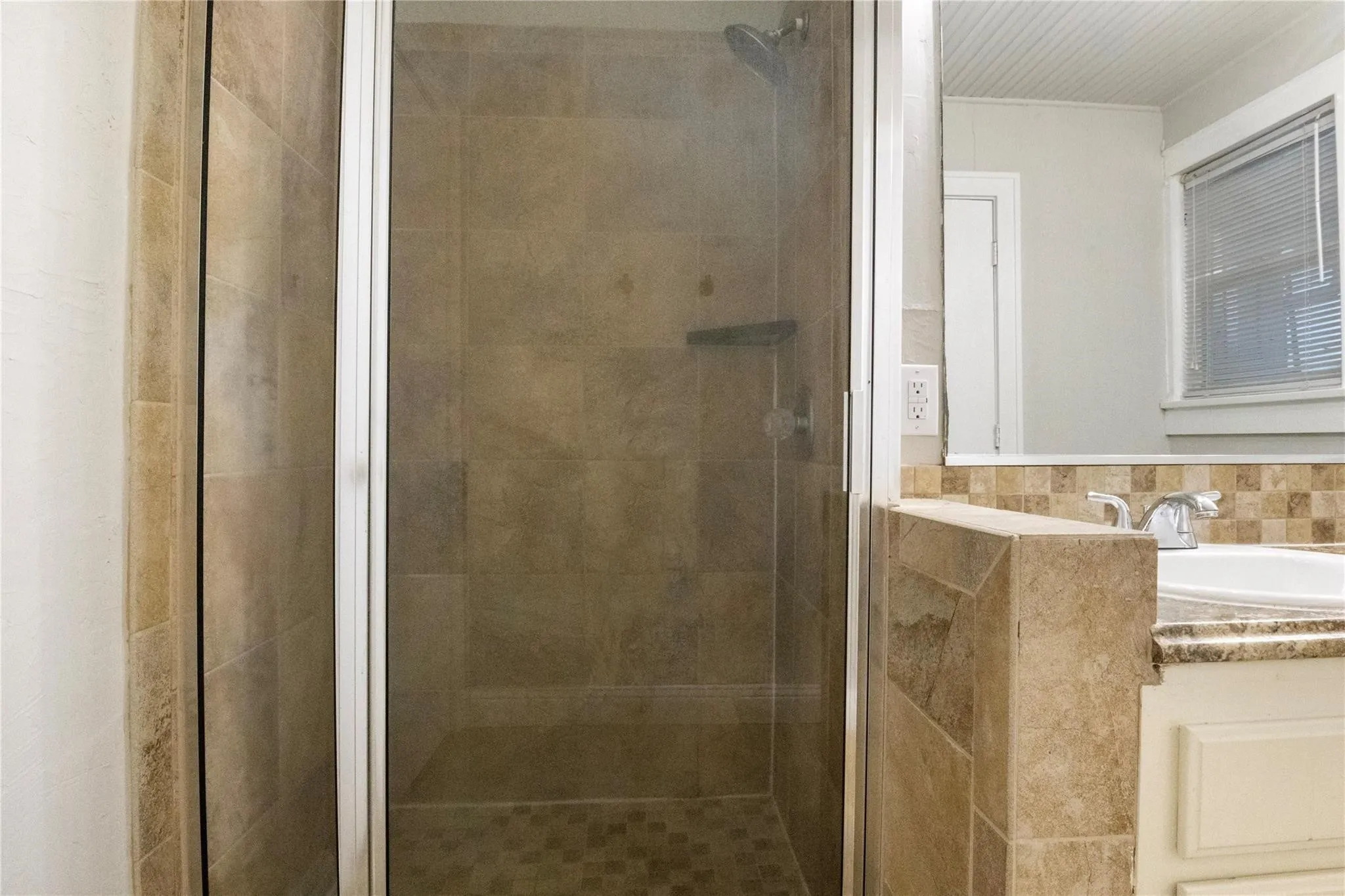 Walk-in shower with full-height tile surround, glass-paneled door, and integrated shelving, featuring a vanity with a stone-finish countertop, undermount sink, and mosaic tile backsplash