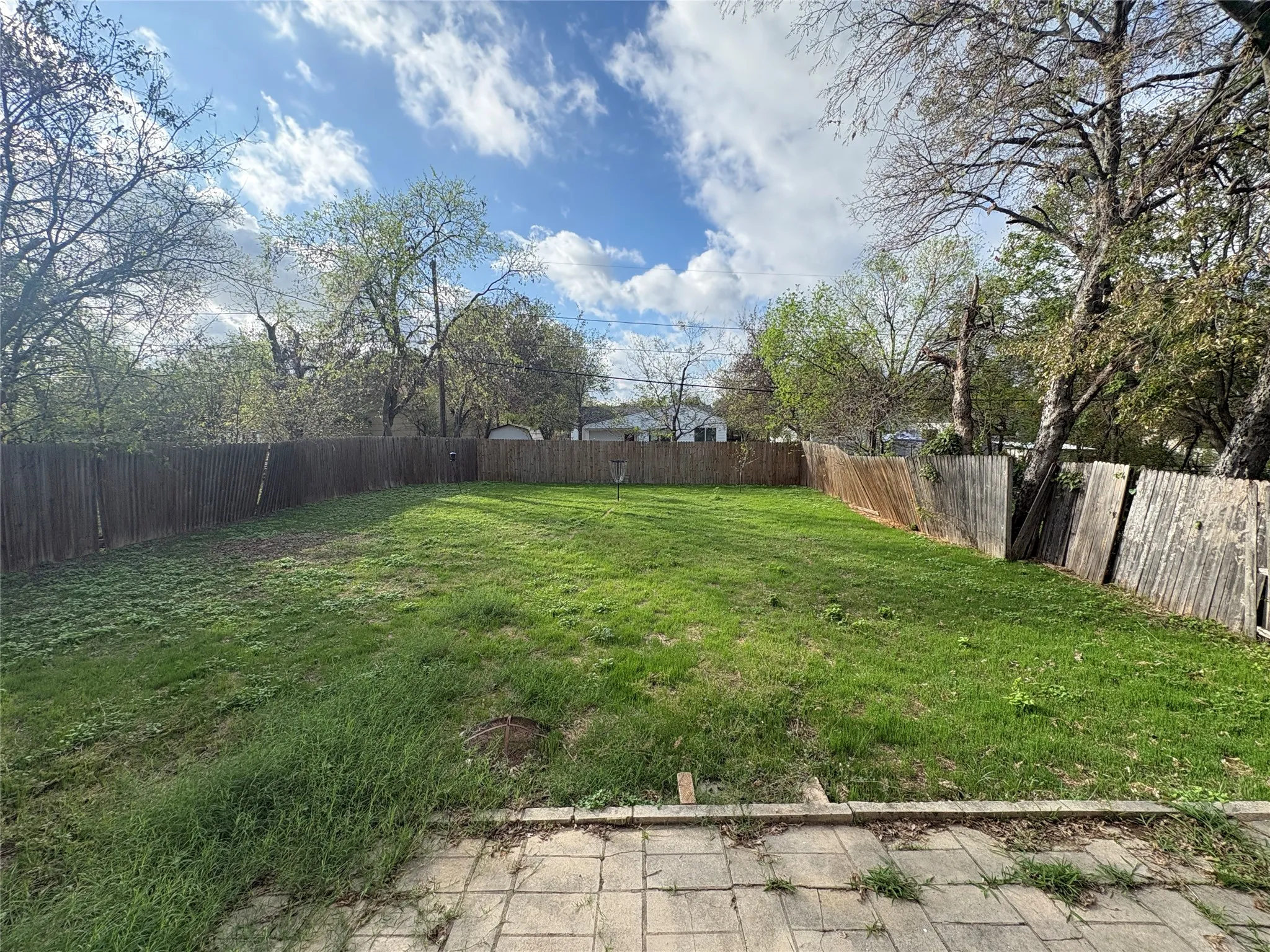 Rear yard featuring a grass lawn, paved stone patio, wood-slat perimeter fencing, and mature trees