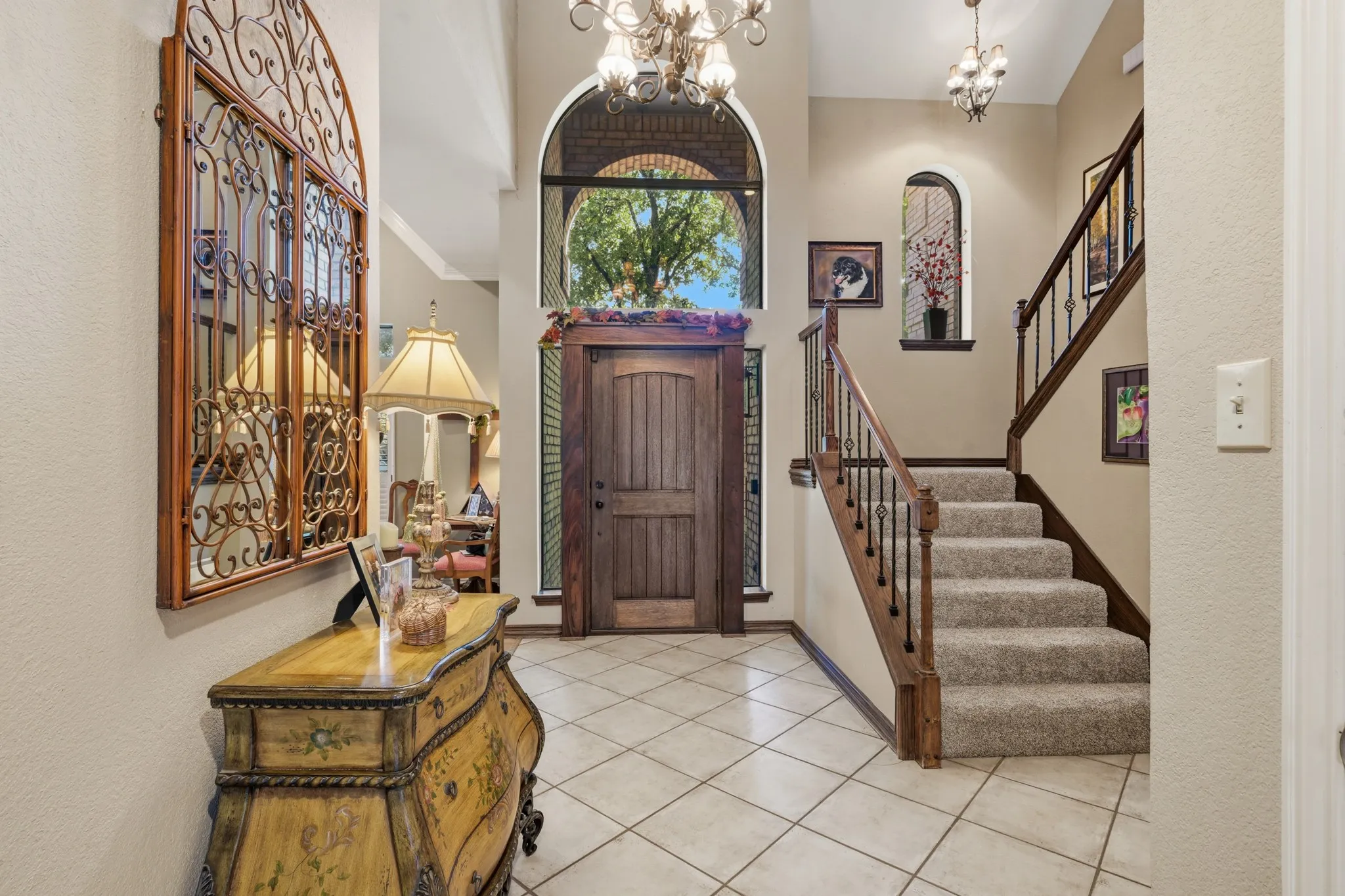 Welcoming entryway featuring a striking wood front door framed by an elegant arched window with decorative brick detailing. Soaring ceilings, statement chandeliers, and a beautiful staircase with rich wood handrails and wrought-iron balusters create an impressive first impression.