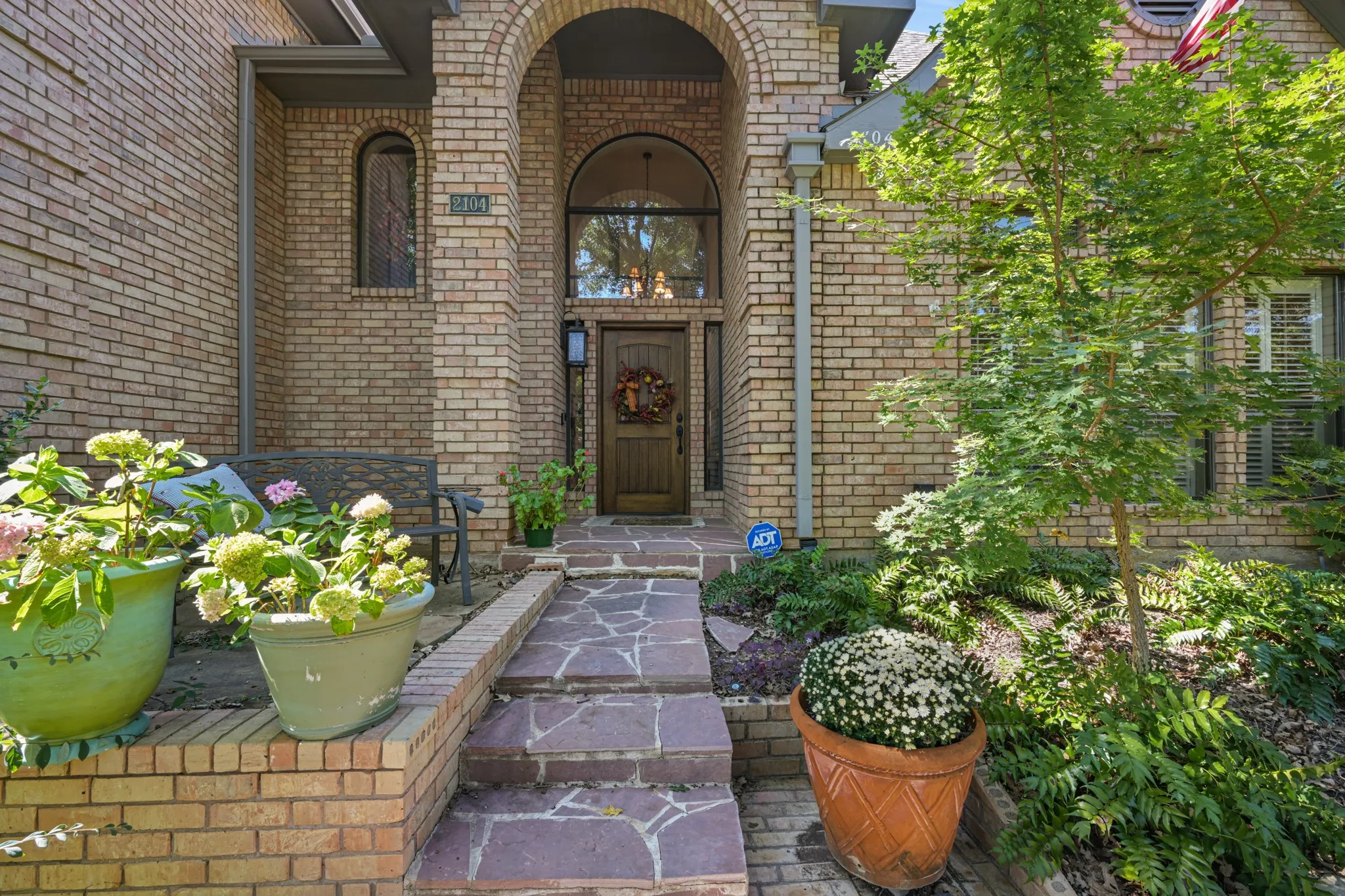 Welcoming brick entryway featuring a charming arched portico, decorative glass above the wood front door, and a flagstone walkway leading to the entrance. Mature landscaping, lush greenery, colorful flowering plants, and a cozy seating area create inviting curb appeal and a warm first impression.