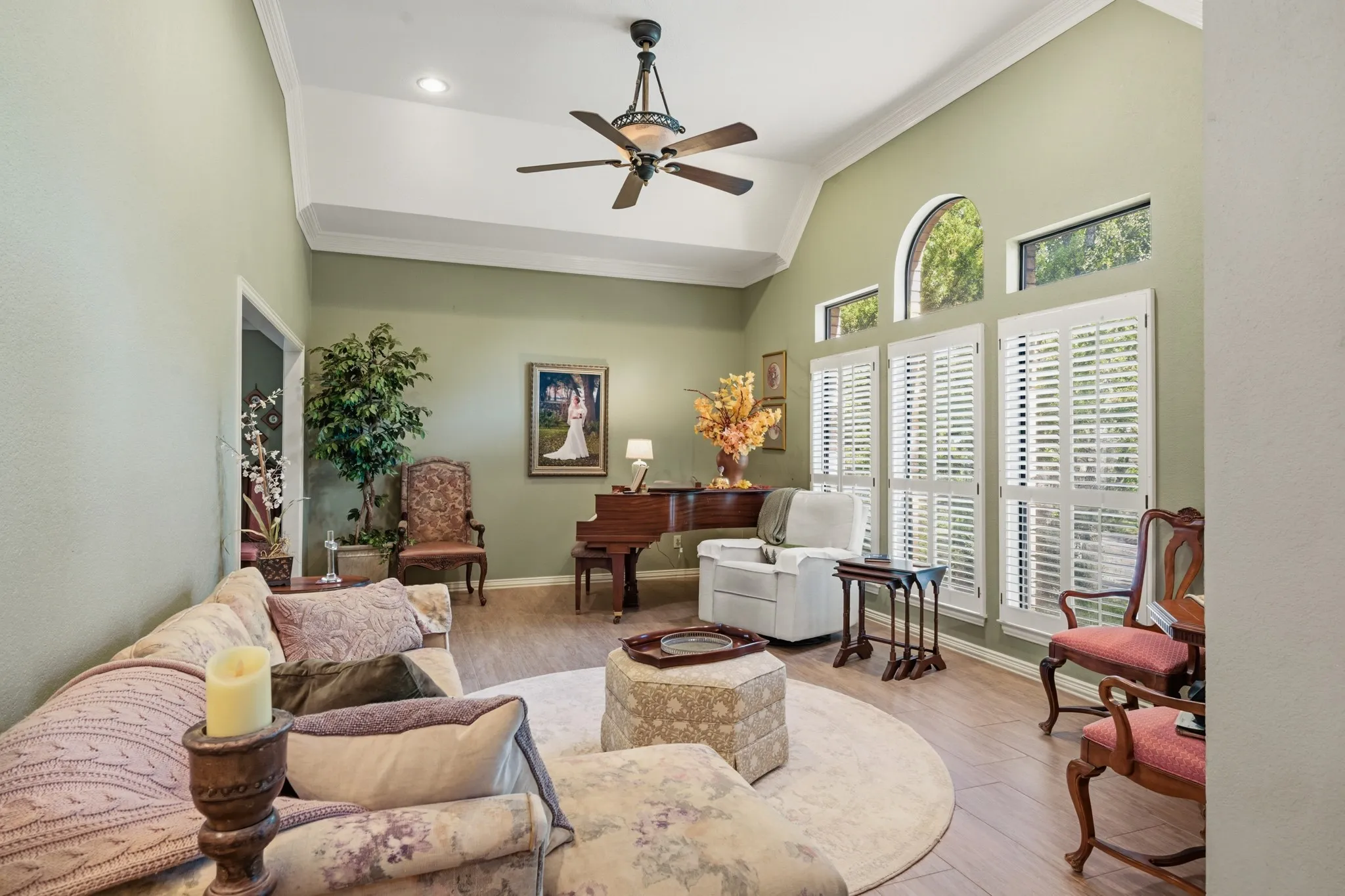 Beautiful formal living room featuring a vaulted ceiling, elegant crown molding, and a ceiling fan. Expansive windows with plantation shutters and an arched transom window fill the space with natural light.