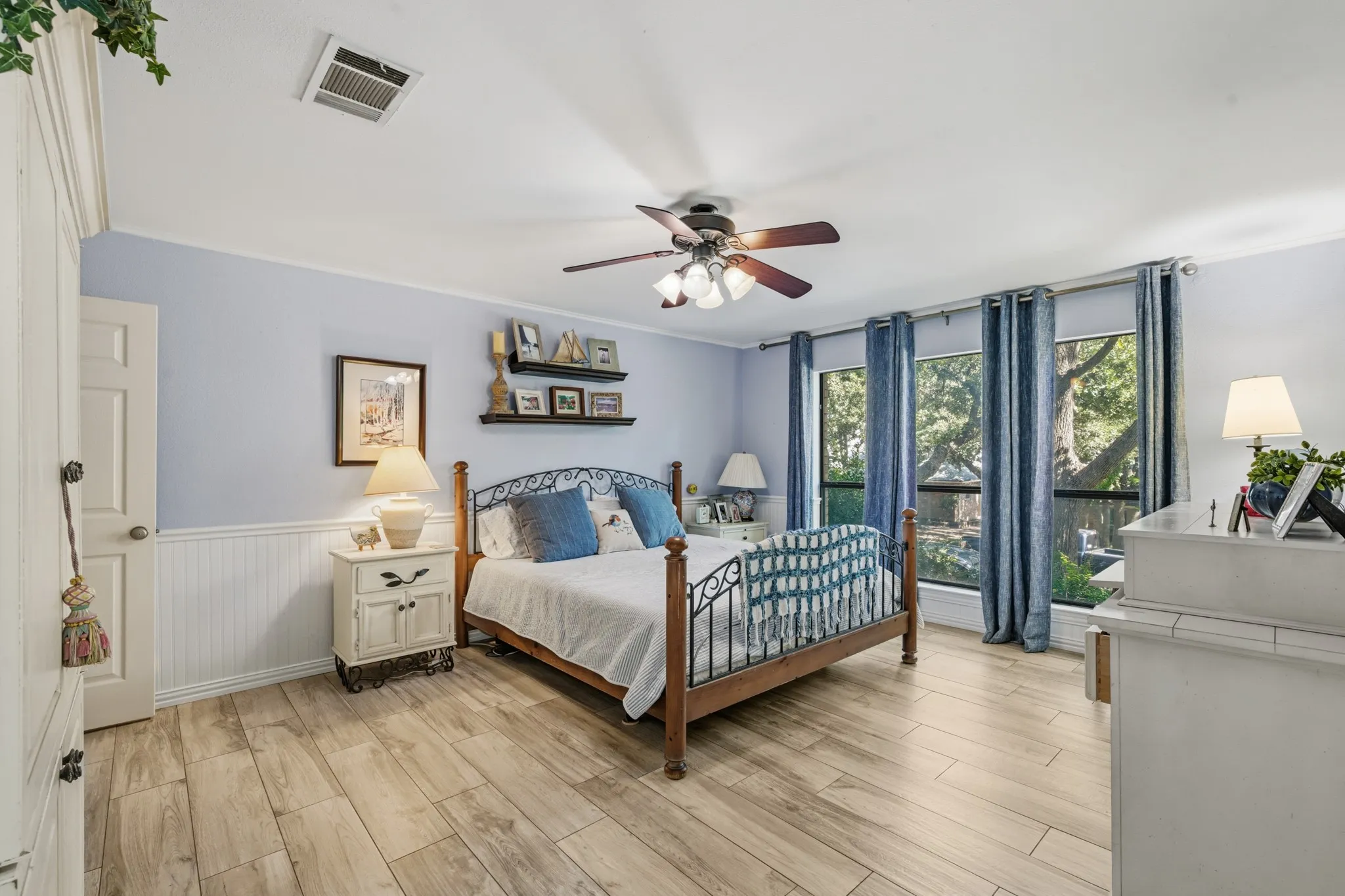 Spacious primary bedroom featuring light wood-look flooring, soft blue walls, and elegant white wainscoting.
