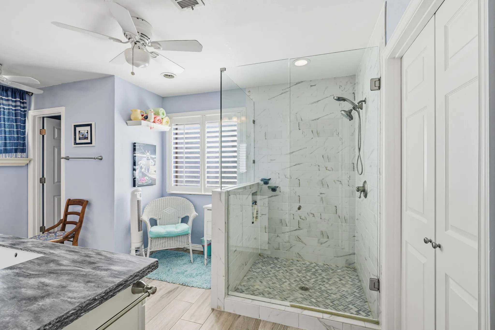Spacious primary bathroom featuring a large walk-in shower with elegant marble-look tile, decorative mosaic flooring, and sleek frameless glass enclosure. Light wood-look flooring, soft blue walls, and crisp white cabinetry create a bright, refreshing atmosphere.