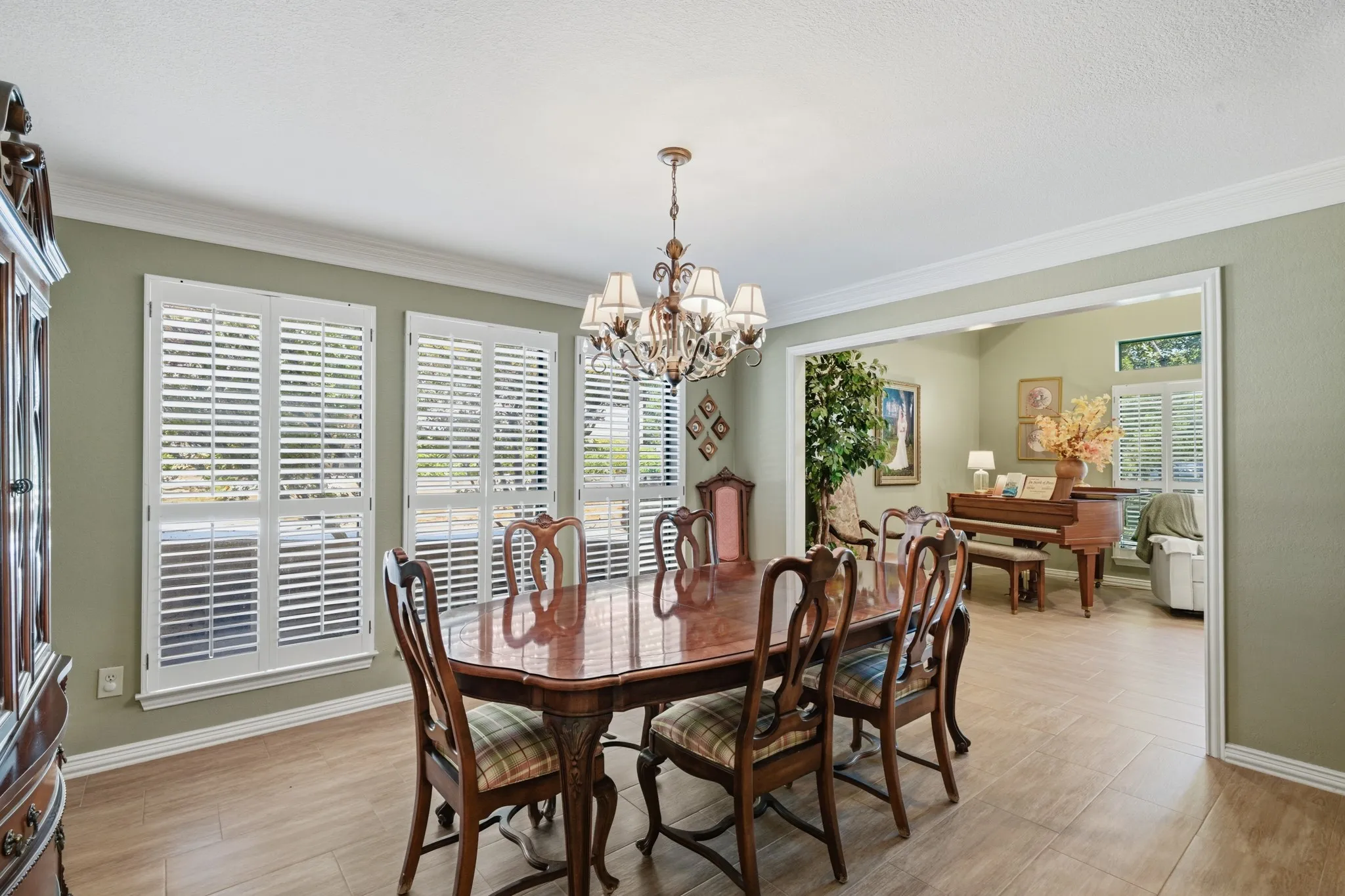 Formal dining room features warm wood-look flooring, decorative crown molding, and soft neutral-green walls create a timeless atmosphere. Plantation shutters span the windows, providing abundant natural light and a beautiful backdrop for entertaining and special gatherings.