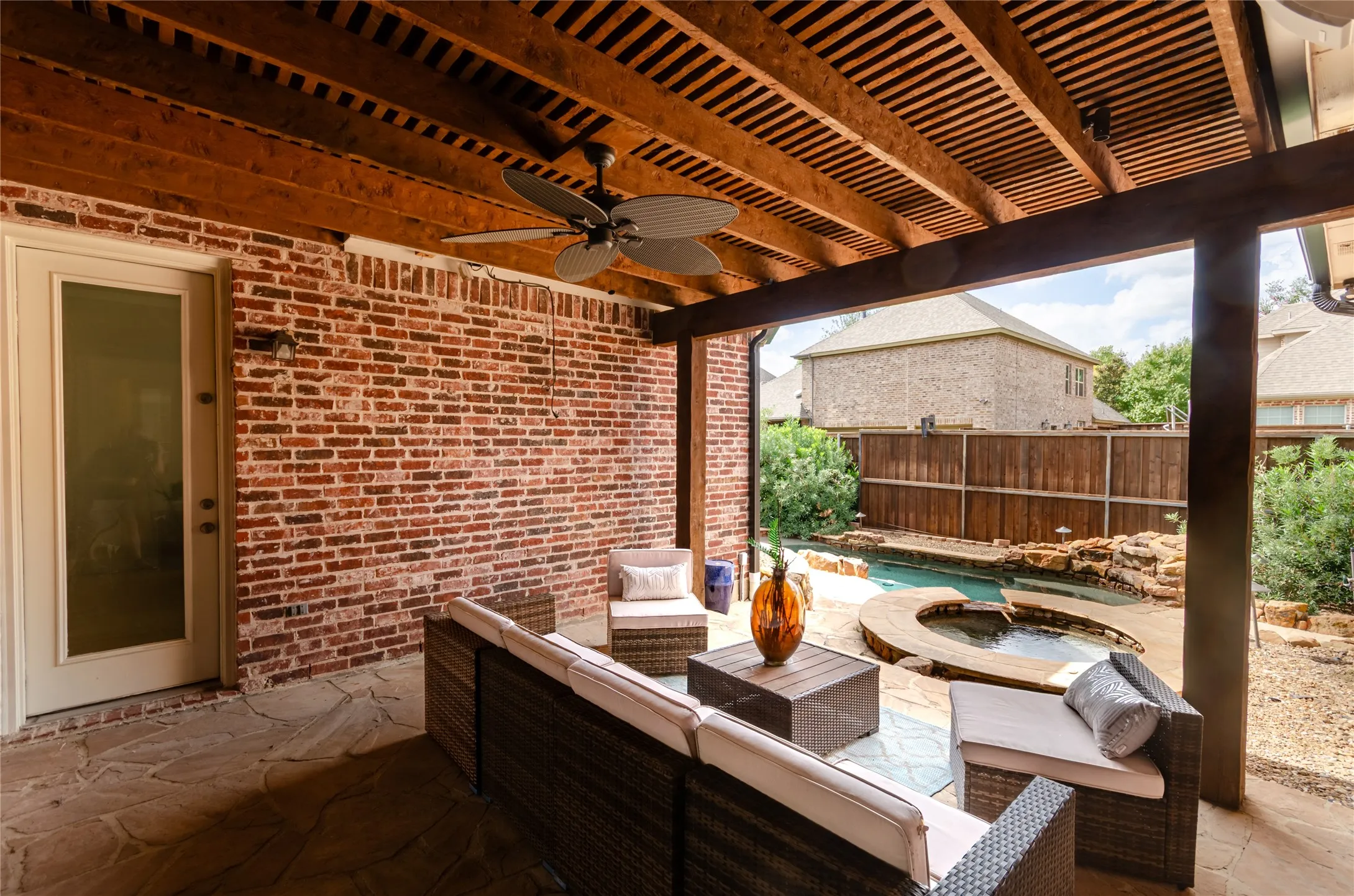 Covered outdoor patio featuring a slatted wood ceiling, exposed brick wall, stone-paved flooring, built-in ceiling fan, and access to a swimming pool with a stone-surround spa
