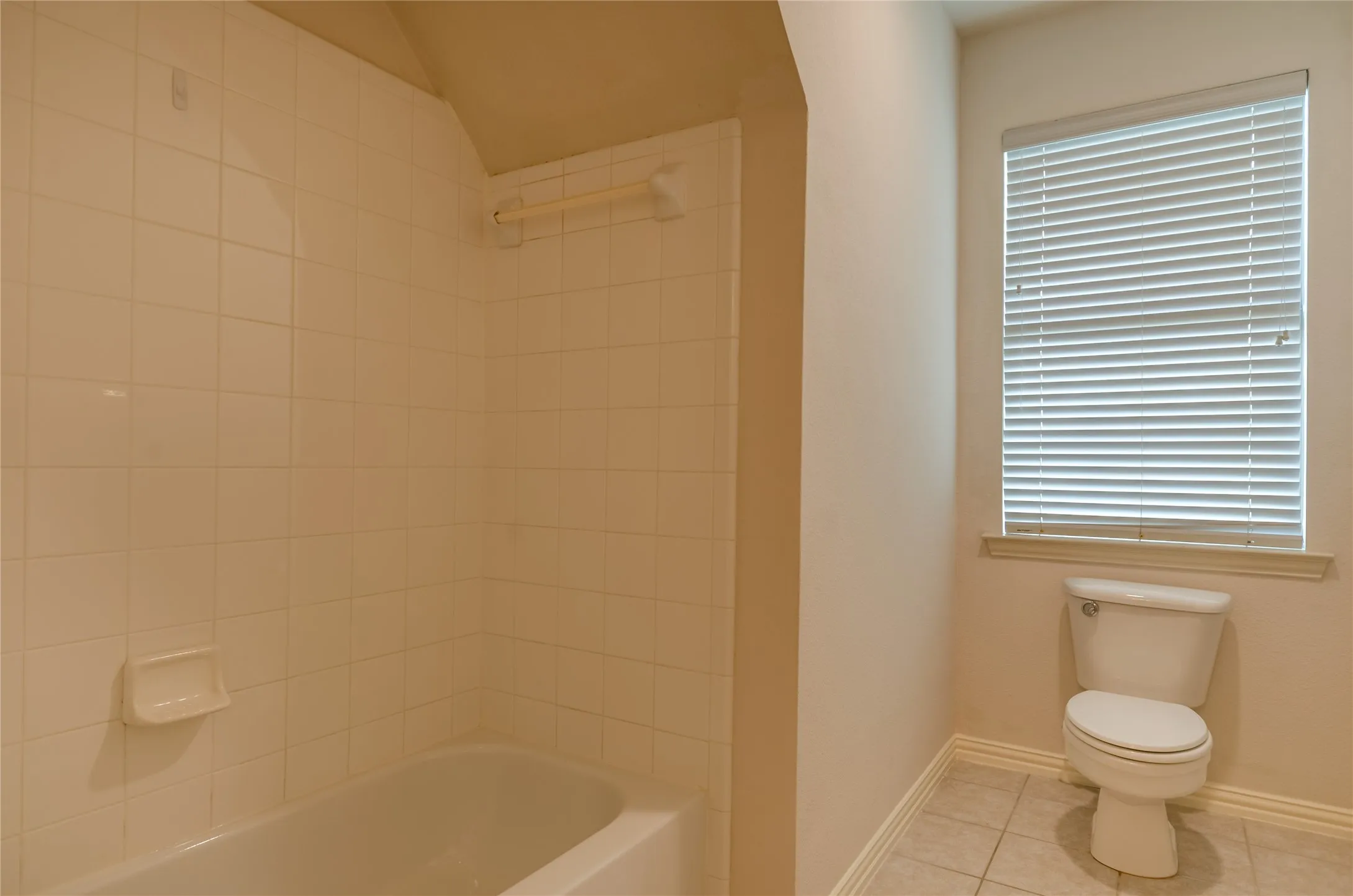 Bathroom featuring a porcelain tub with white tile surround, built-in soap dish, integrated grab bar, two-piece toilet, and window with horizontal blinds