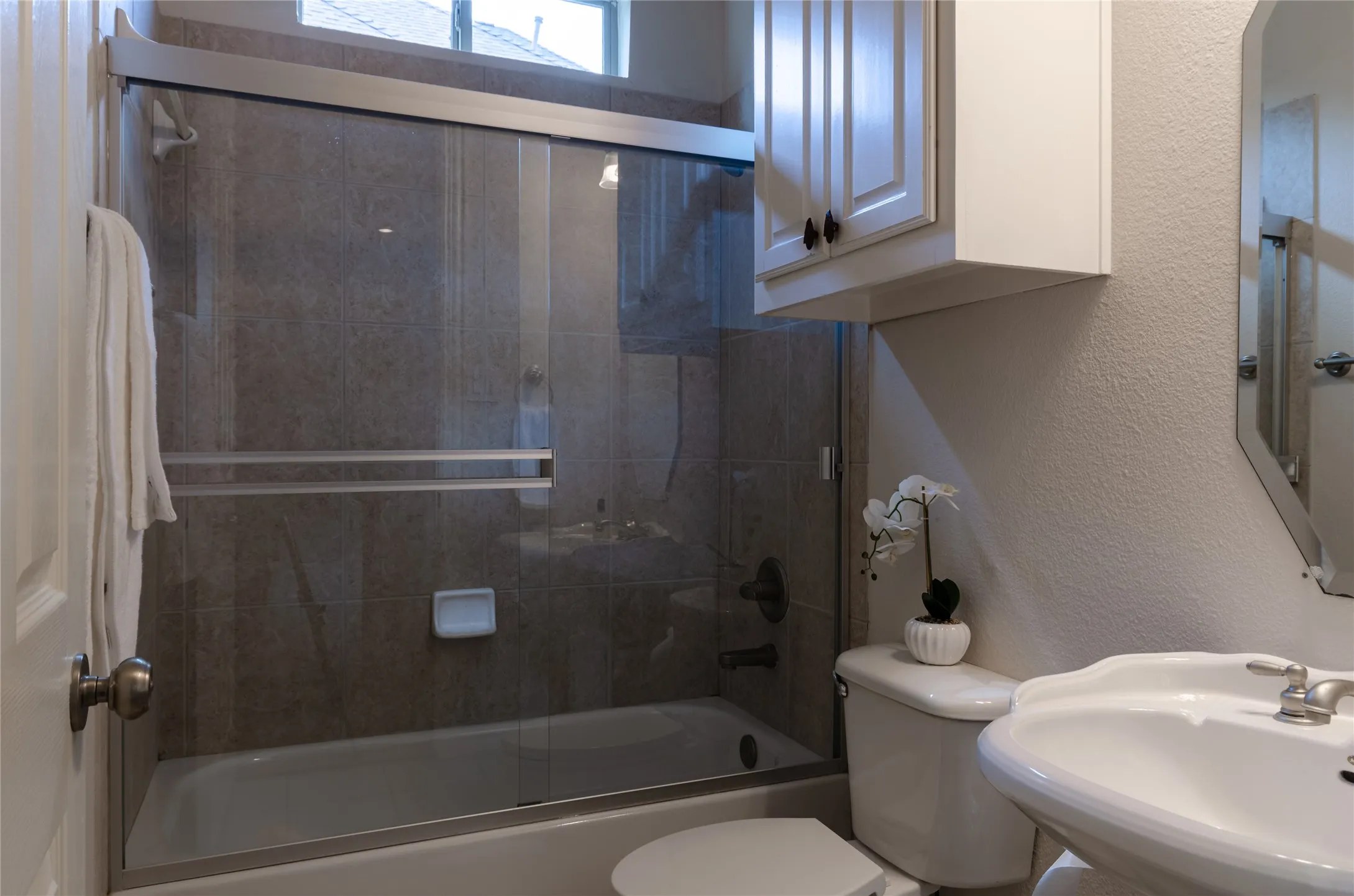 Full-height tiled shower surround, sliding glass enclosure, white upper wall cabinet, and pedestal sink with metallic fixtures