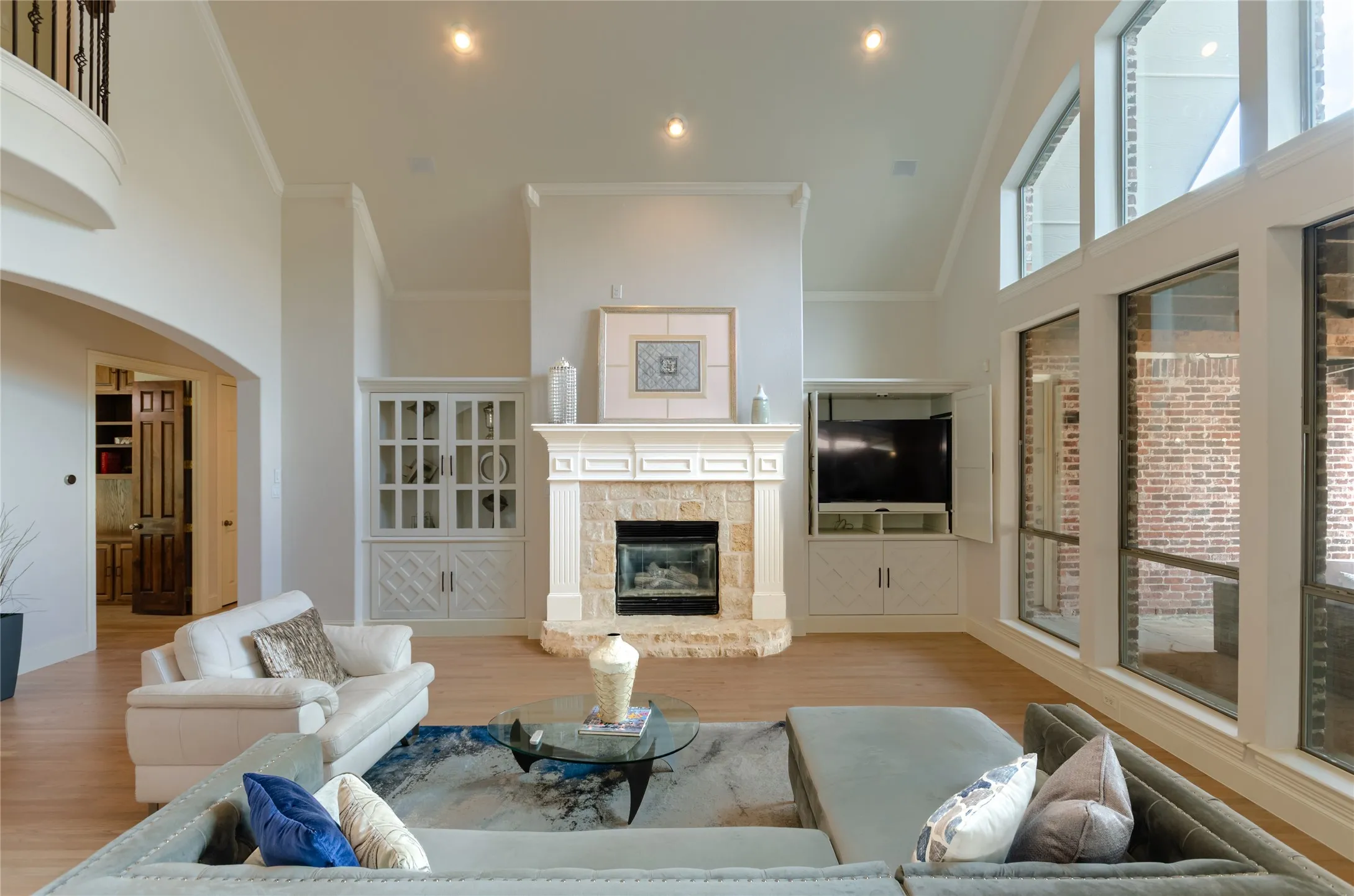 Vaulted ceilings, floor-to-ceiling windows, stone-surround fireplace with a custom mantle, and built-in cabinetry featuring glass-front display shelving and hidden media storage