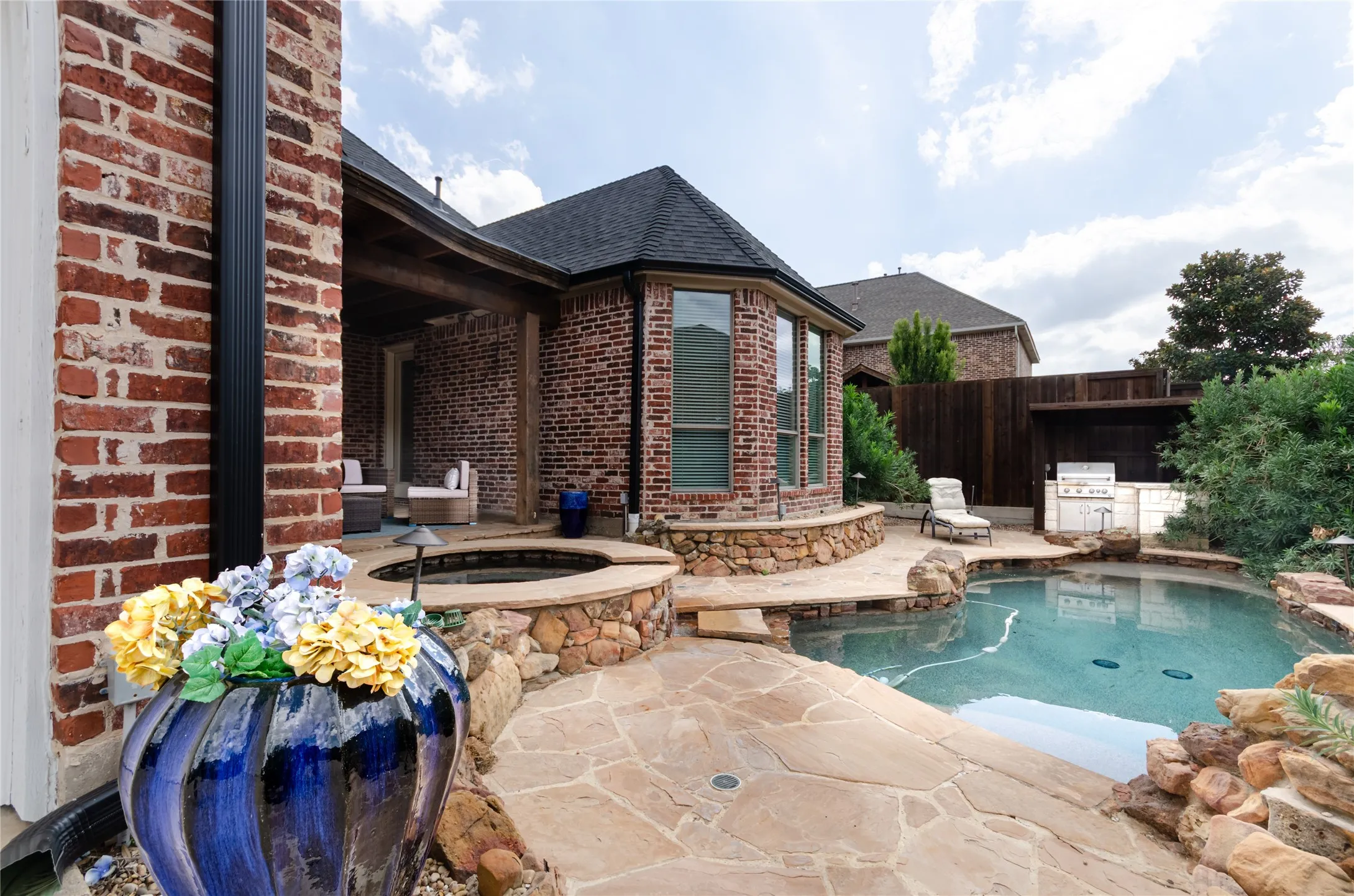 Brick exterior featuring a covered patio, stone-encased spa, custom swimming pool with rock accents, and built-in outdoor cooking station