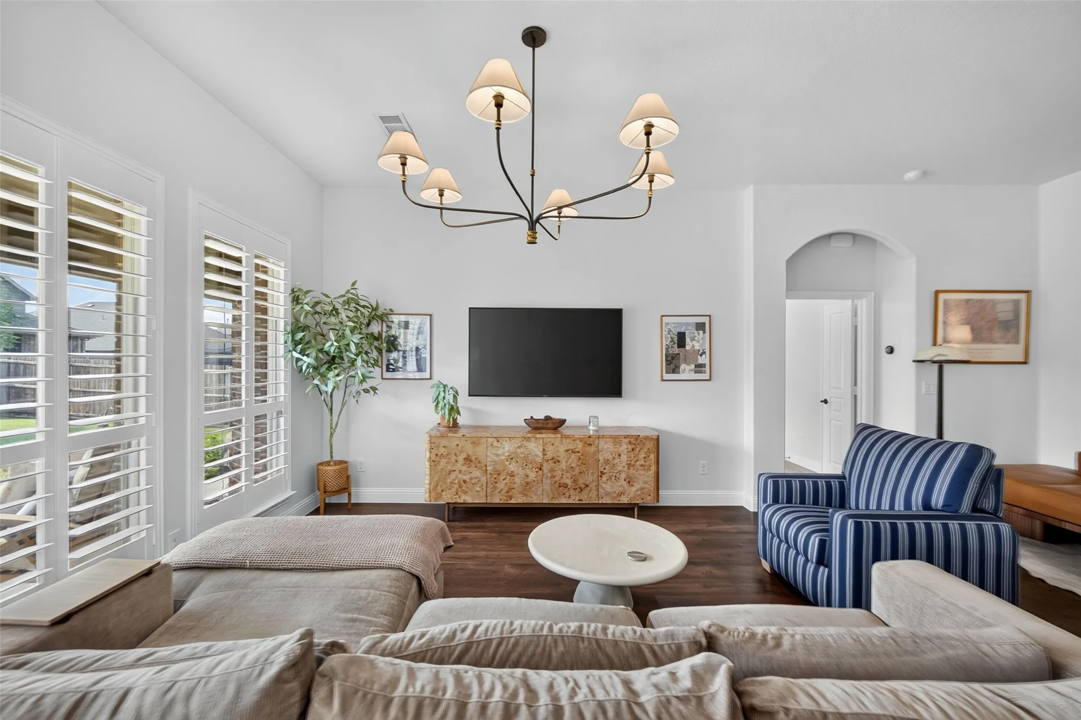 Living area featuring dark wood-finish flooring, six-arm chandelier with fabric shades, floor-to-ceiling shutters, and an arched entry