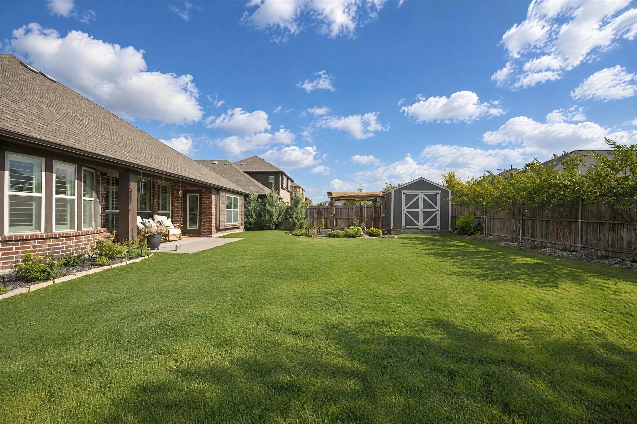 Expansive backyard featuring a concrete patio, manicured lawn, wooden perimeter fencing, detached storage shed, and a pergola