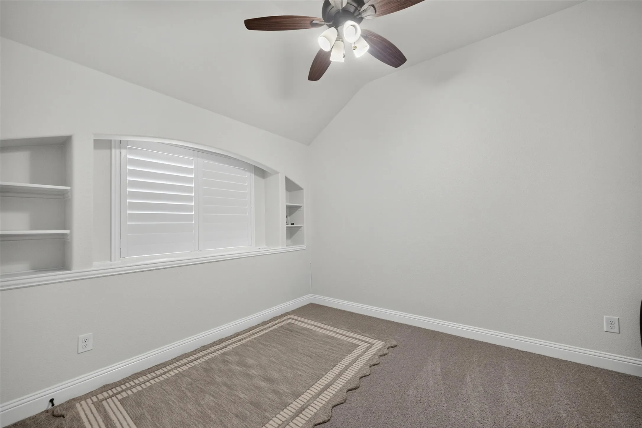 Vaulted ceiling featuring a central multi-blade ceiling fan, arched window with plantation shutters, and symmetrical built-in shelving