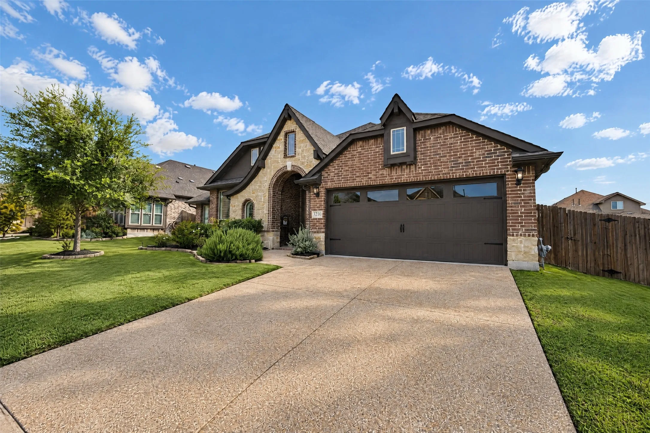 Exterior elevation featuring a combination of brick and stone veneer, a multi-gabled roofline, a multi-car garage with glass panels, and an aggregate concrete driveway