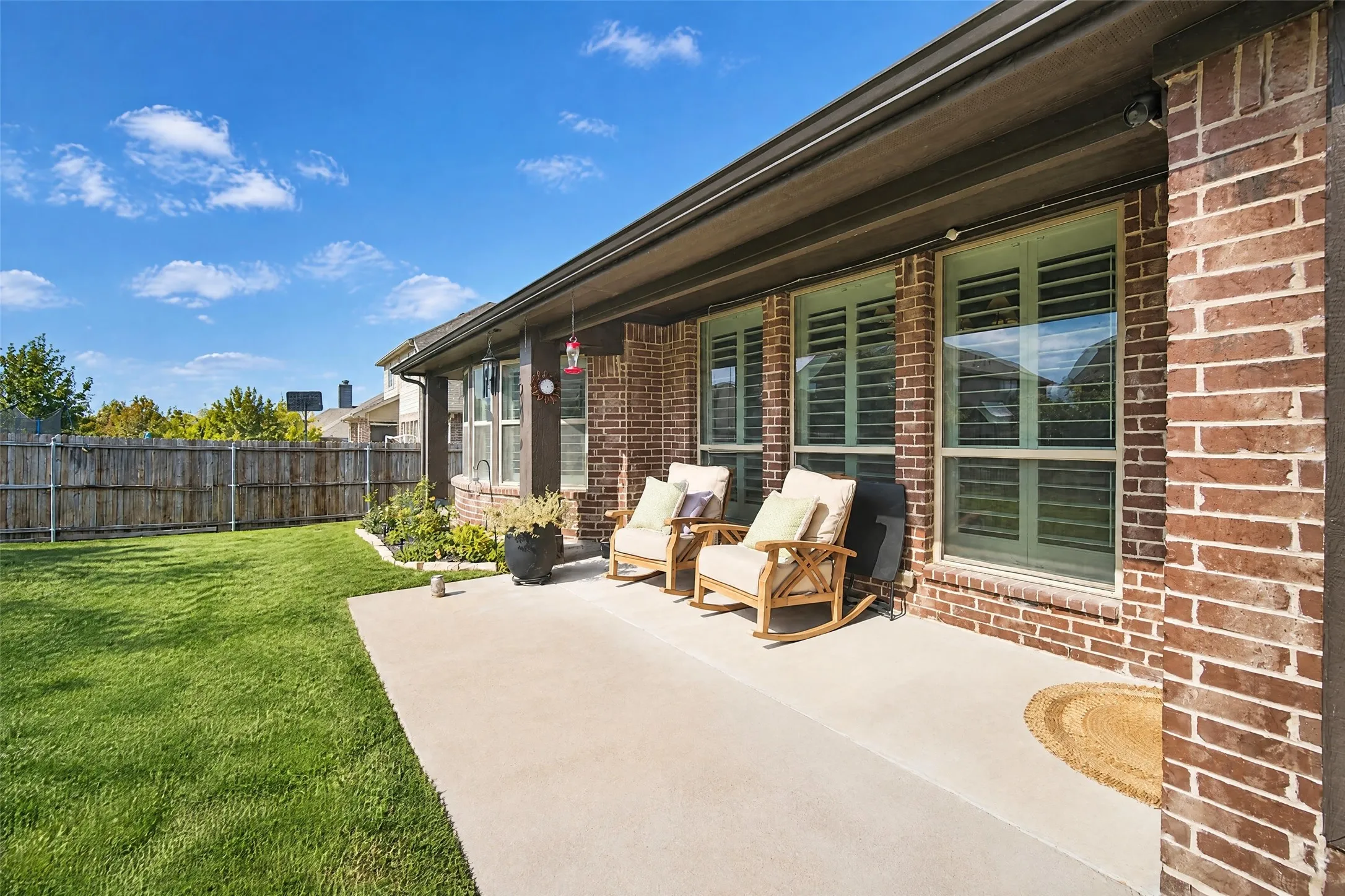 Covered concrete patio featuring brick exterior siding, plantation-style window shutters, and a fenced backyard with lawn