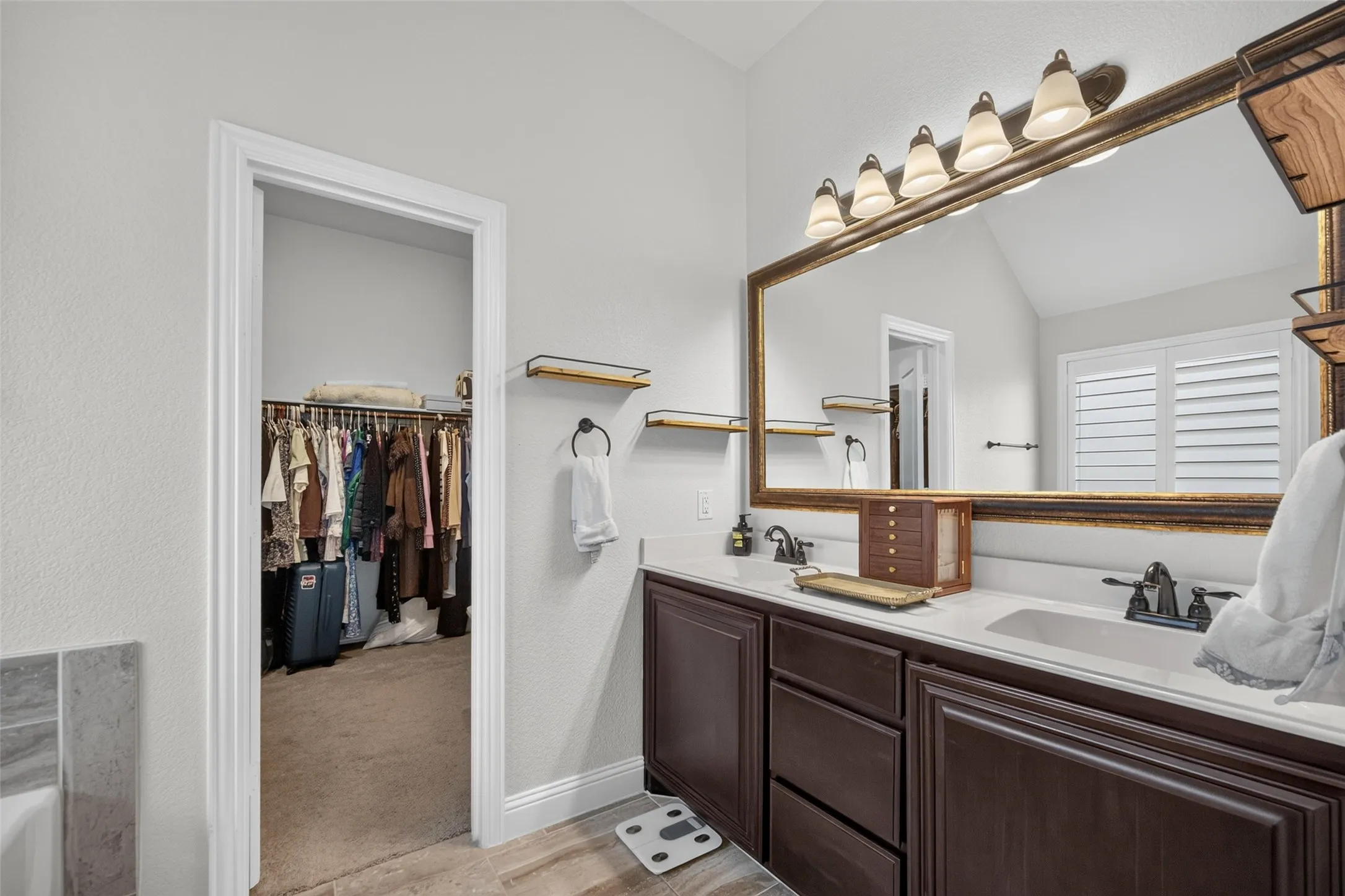 Double-sink vanity with dark-finish cabinetry, solid-surface countertop, dual metal faucets, and a large framed vanity mirror with integrated multi-bulb lighting
