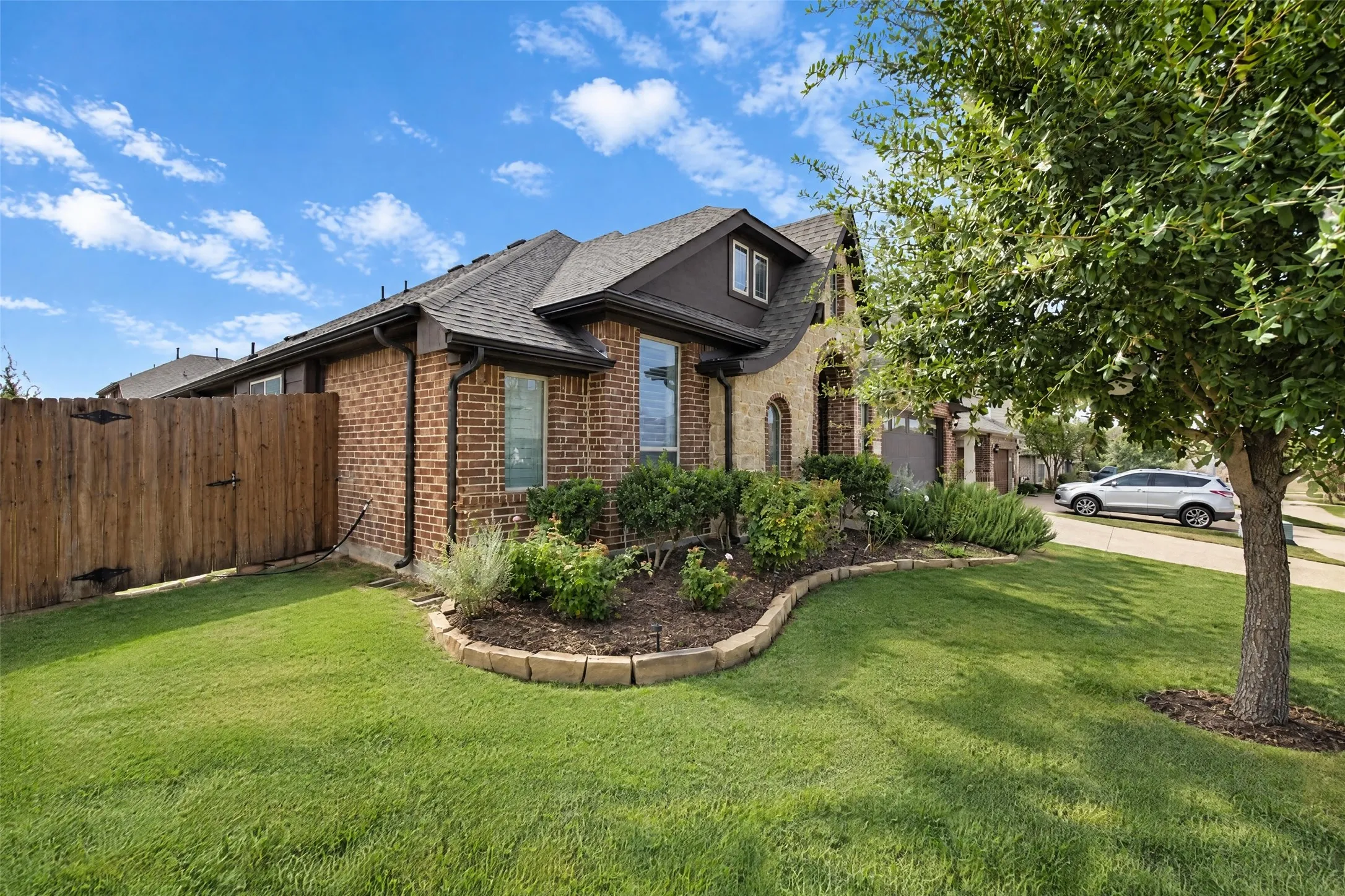 Brick and stone veneer exterior featuring a gabled roofline, manicured lawn, stone-bordered landscape beds, and wood privacy fencing