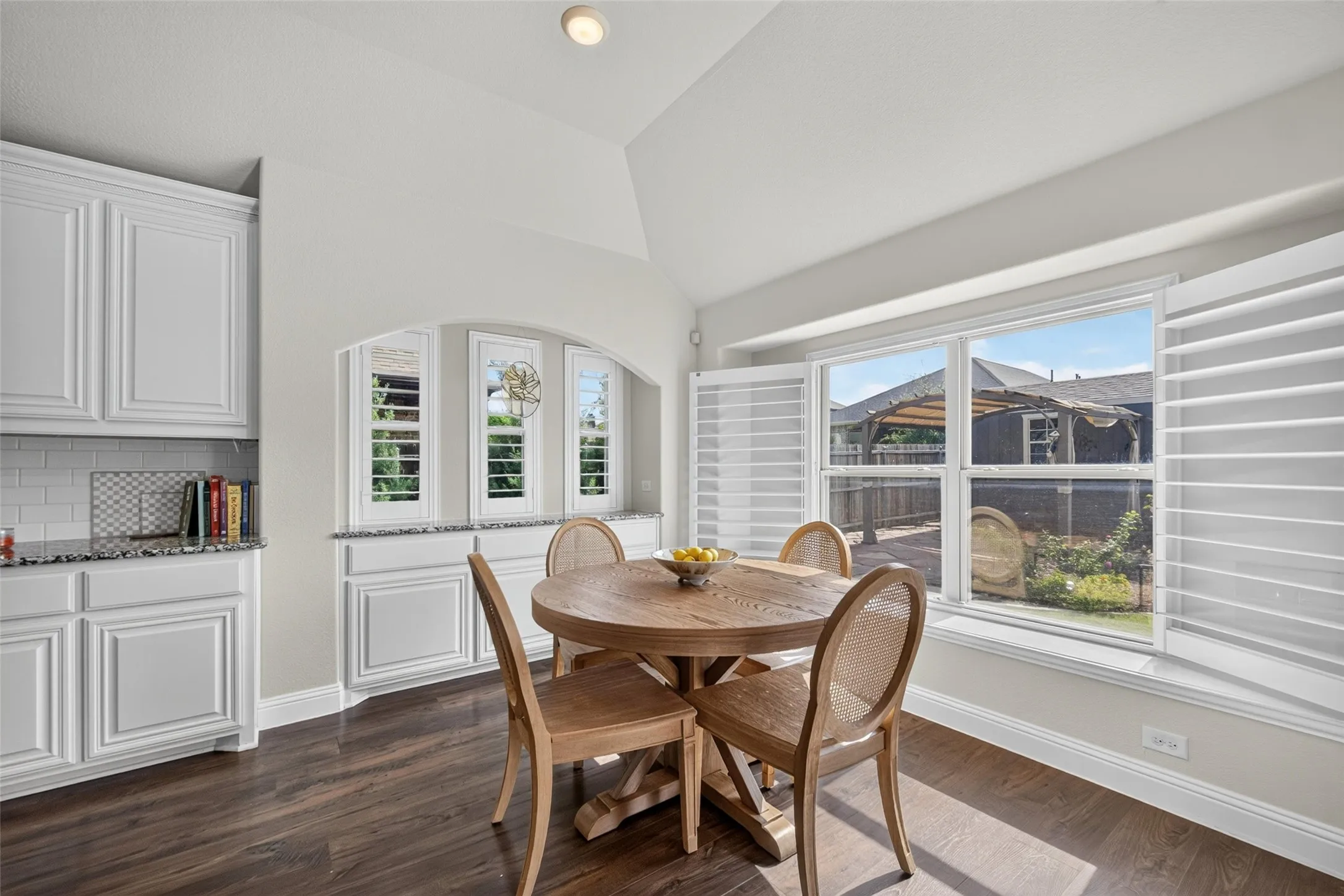 Vaulted ceiling, white shaker-style cabinetry with stone countertops, built-in recessed cabinetry with arched alcove, wood-finish flooring, and large windows featuring plantation shutters