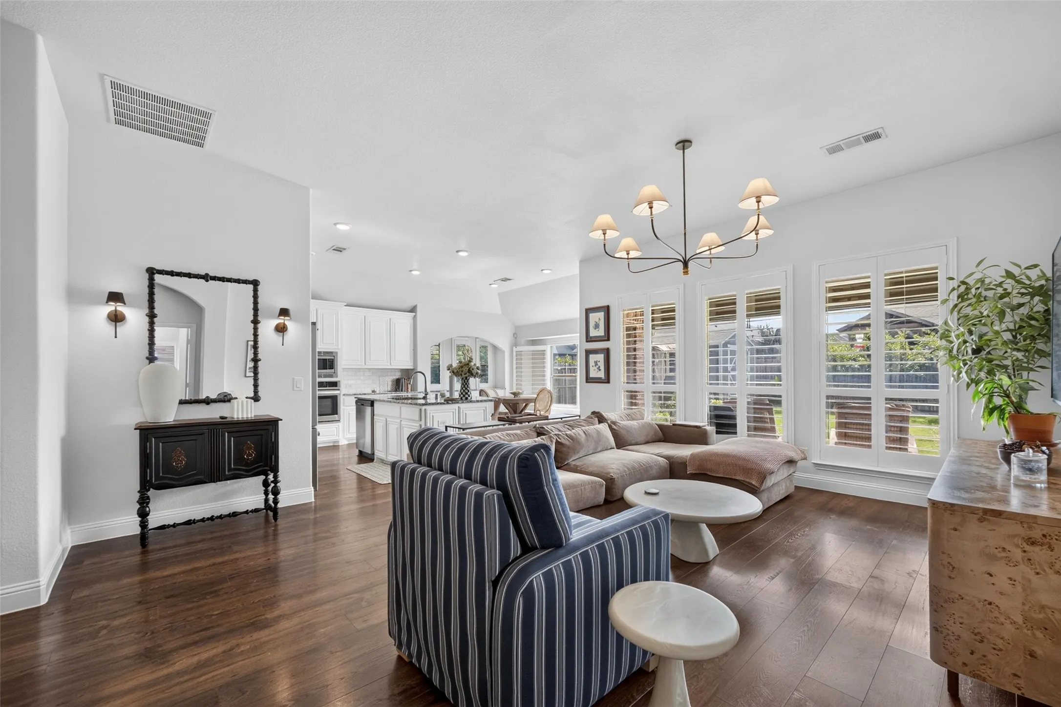 Open-concept interior featuring wood-finish flooring, multi-pane windows with plantation shutters, vaulted ceilings, and a decorative chandelier
