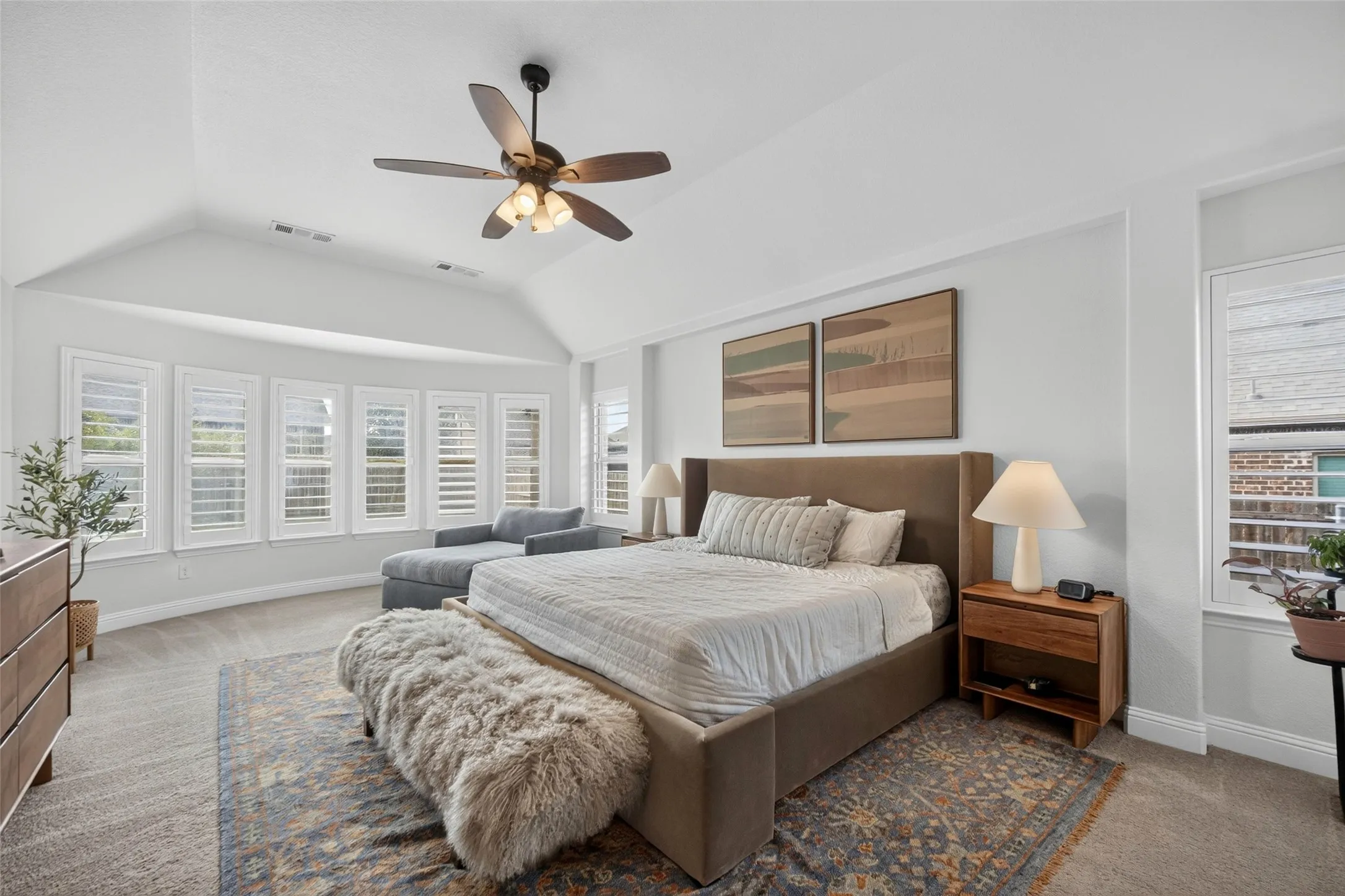 Vaulted ceiling with a central multi-blade ceiling fan, expansive bay window featuring plantation shutters, and neutral carpeting
