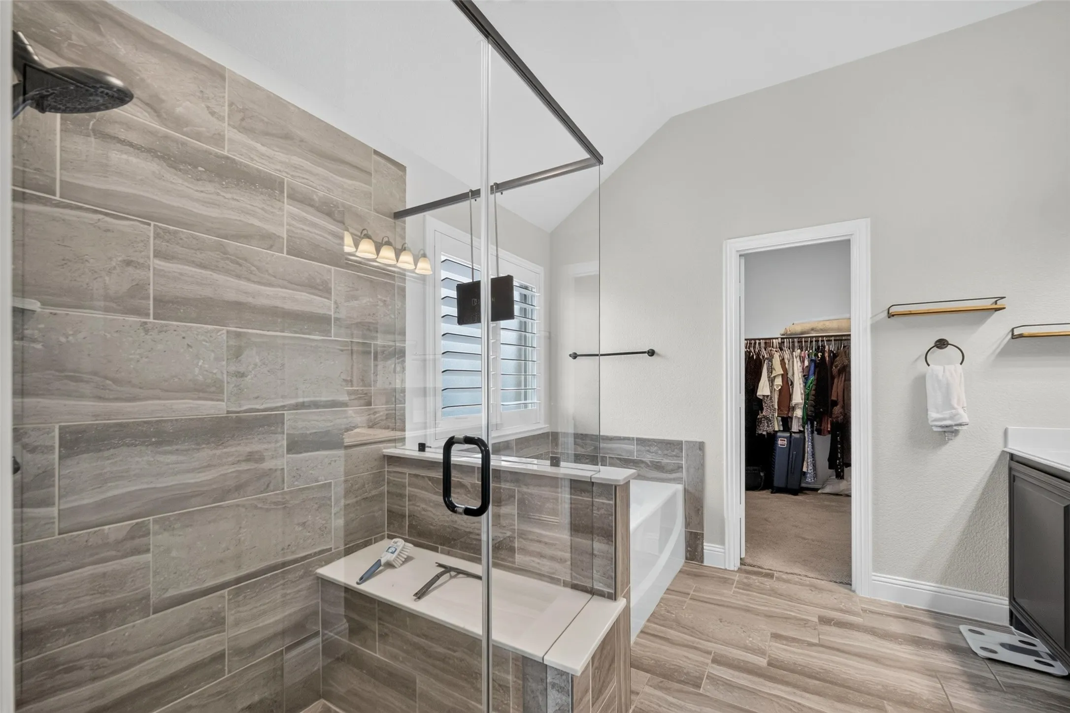 Walk-in shower featuring glass paneling, floor-to-ceiling stone-finish tile, and built-in bench seating, complemented by wood-finish floor tiling and a soaking tub